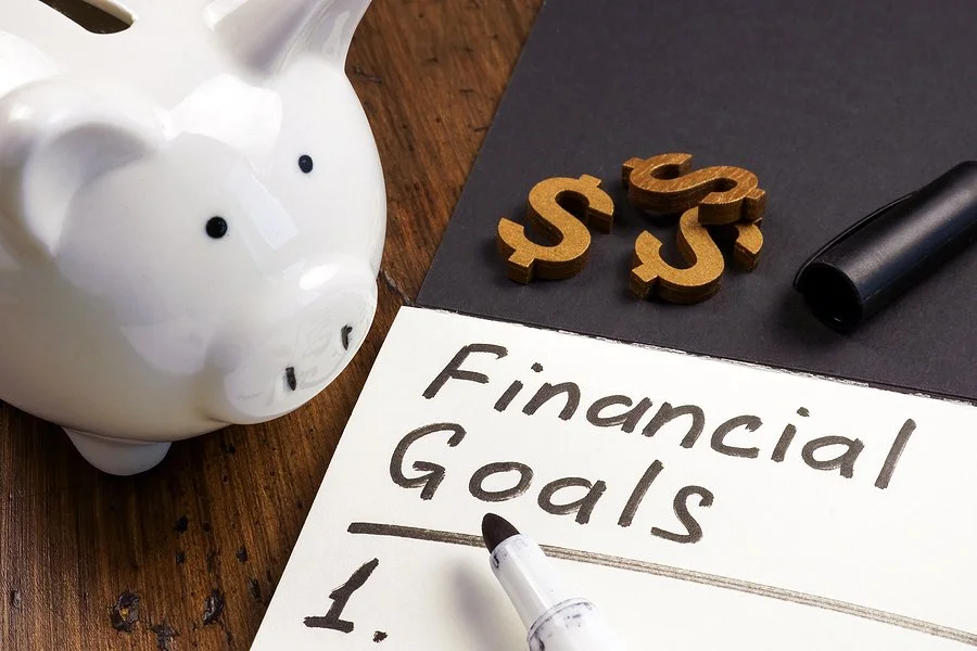 A white piggy bank, wooden dollar sign decorations, a black marker, and a white sheet of paper with 'Financial Goals' written on it.
