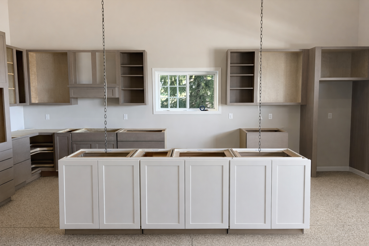 Kitchen cabinets under construction with empty frames, light-colored walls, a central window, and an unfinished island in a new home.