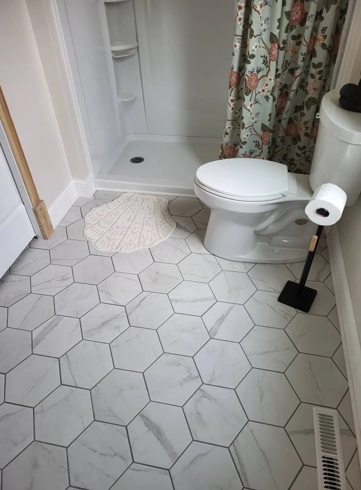 A clean bathroom with white hexagonal marble floor tiles, a white toilet with a toilet paper roll on a black stand nearby, a small beige rug in front of the shower, a floral shower curtain, and a white shower stall with shelves.