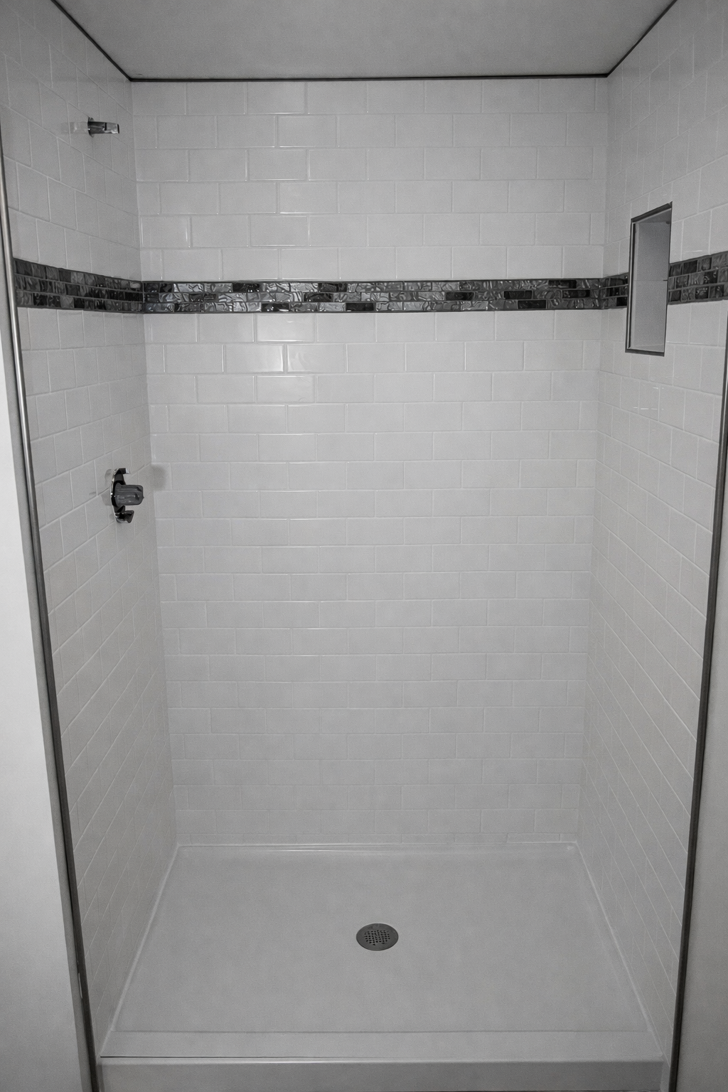 Empty shower with white tiled walls, a decorative black tile border, a small niche, and a drain on the floor.
