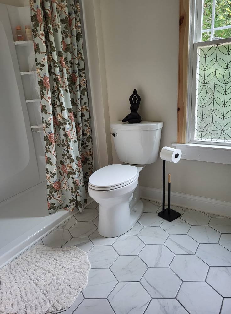 Bathroom with white toilet, black sculpture on toilet tank, patterned shower curtain, window with decorative grid, hexagonal white tiles, and a white bath mat.