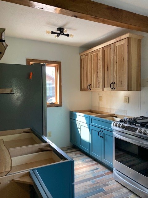 An unfinished kitchen with blue lower cabinets, wooden upper cabinets, a stove, and a small window on the wall.