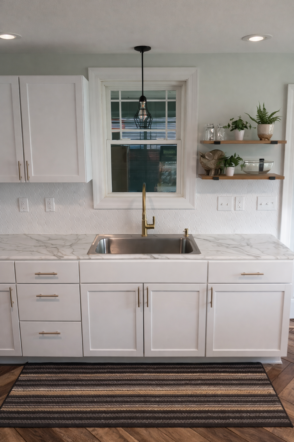 A modern kitchen with white cabinets, a marble countertop, a brass sink faucet, a window with blinds, and wooden shelves with plants and glass jars.