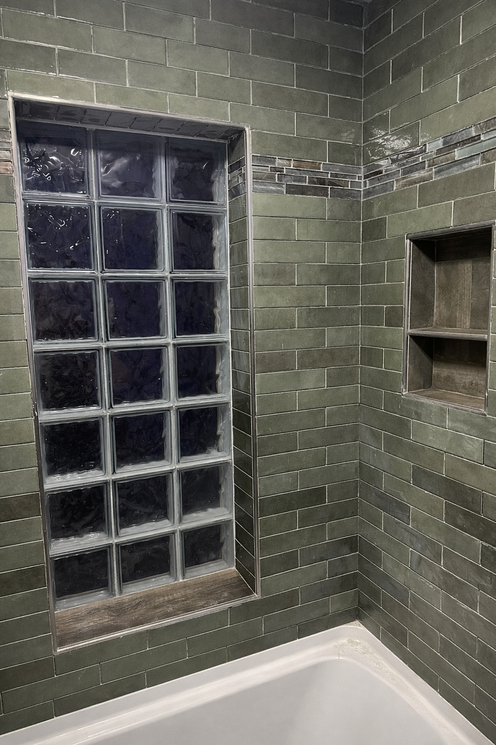 A corner of a bathroom with dark green rectangular tile walls, a built-in shelf niche, and a glass block window.