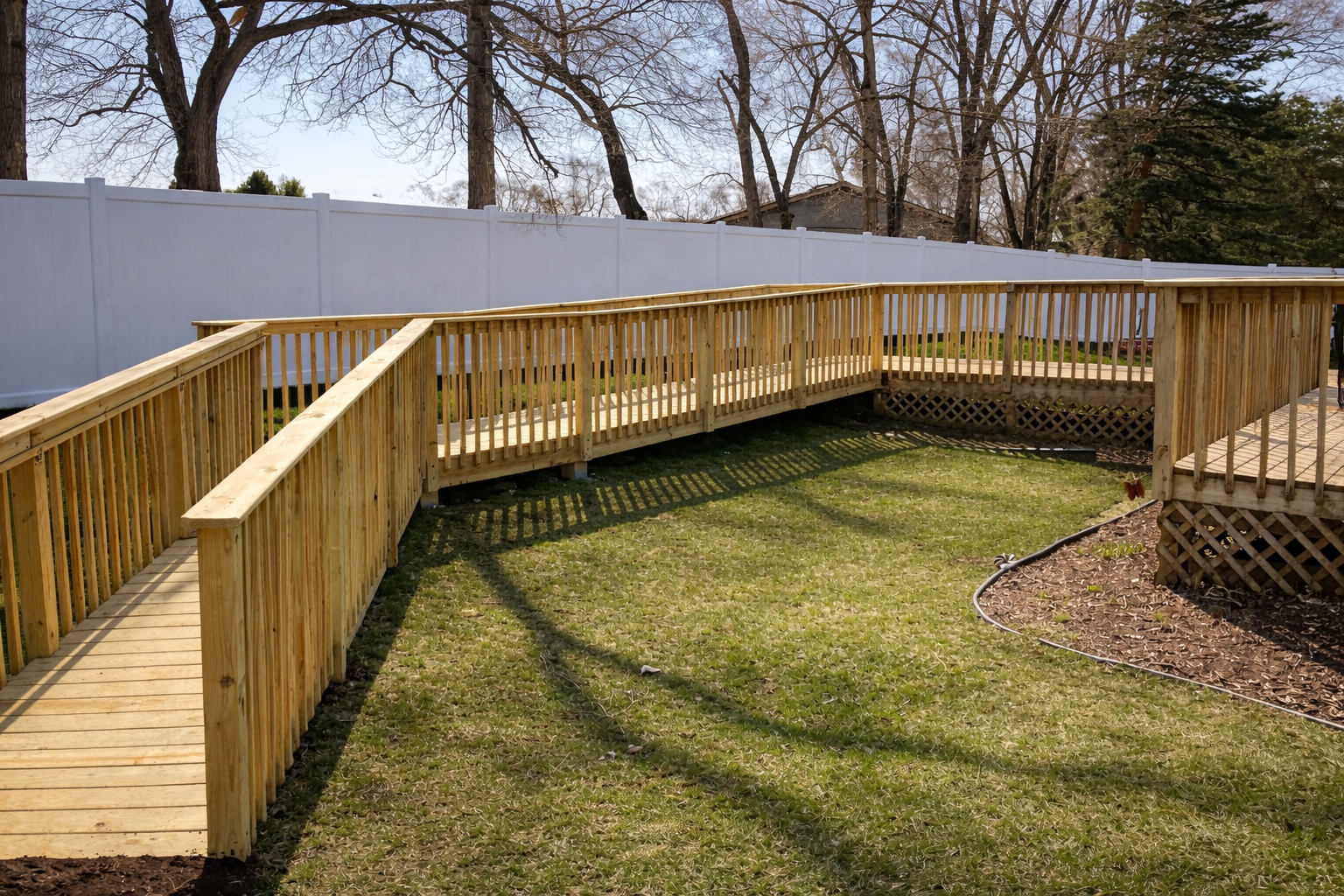 A recently built wooden ramp with railings in a backyard, surrounded by a white fence, with trees and grass.