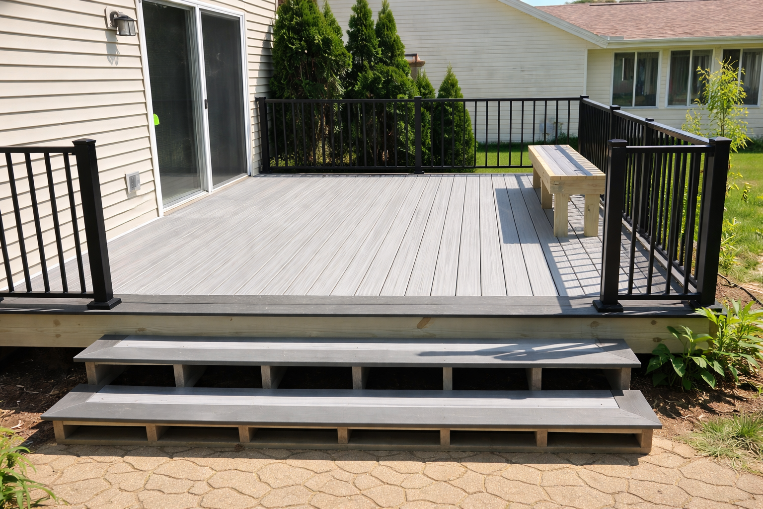 Backyard deck with light gray wooden flooring, black metal railing, and a wooden bench, accessed by three gray steps, with green shrubs and a house with sliding glass door in the background.