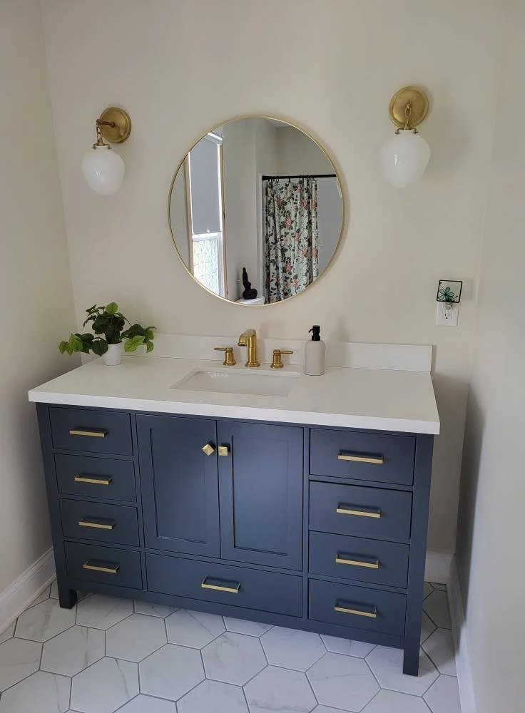 Bathroom vanity with blue cabinetry, white countertop, oval mirror, gold fixtures, two wall sconces, potted plant, soap dispenser, and tiled floor.