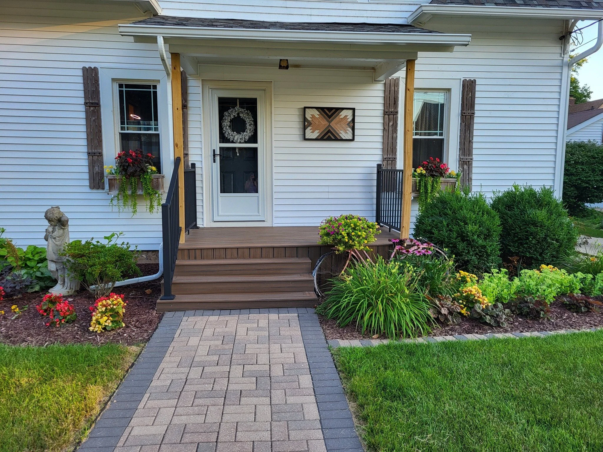 Front porch of a house with flower pots, a statue, a decorated door, and a landscaped garden with plants and bushes.