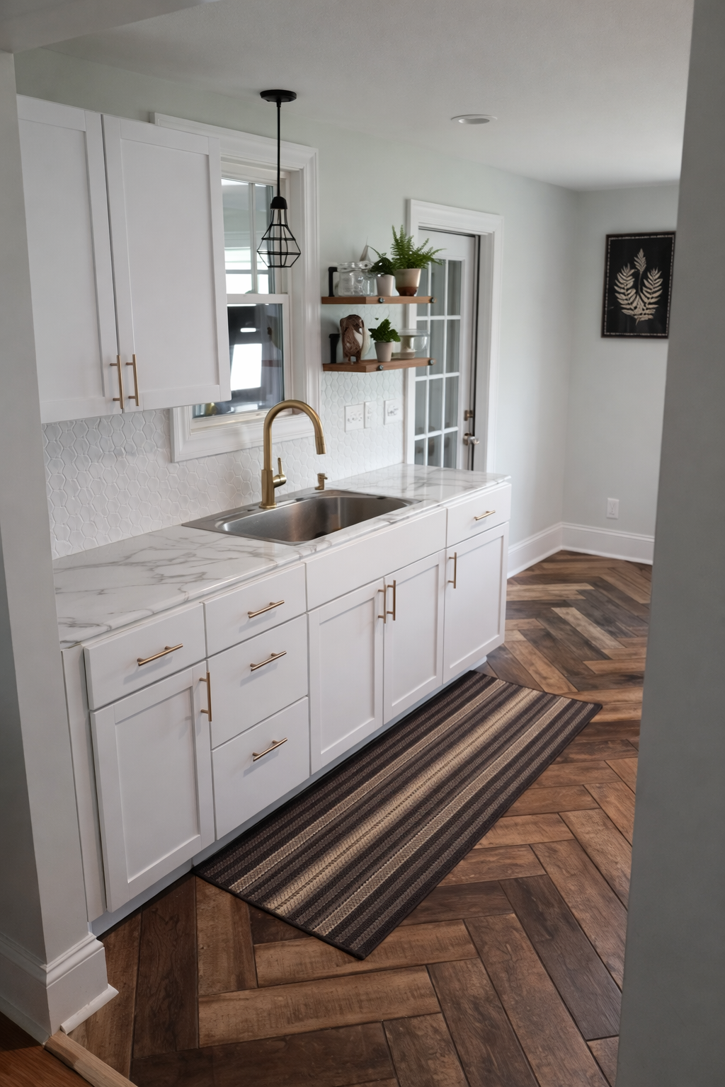 White kitchen with marble countertop, gold handles, gold faucet, white cabinets, and a striped rug on wooden herringbone floor, with a window and decorative shelves with plants and a small sculpture.