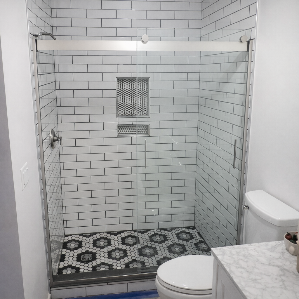 Bathroom with a glass shower enclosure, tiled walls in white subway tiles, and a patterned black-and-white tile floor. A toilet and a marble-topped vanity are visible next to the shower.