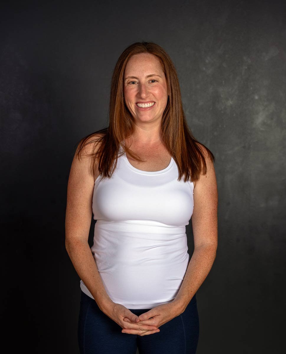 A woman with long red hair, wearing a white sleeveless top, smiling, standing against a dark background.
