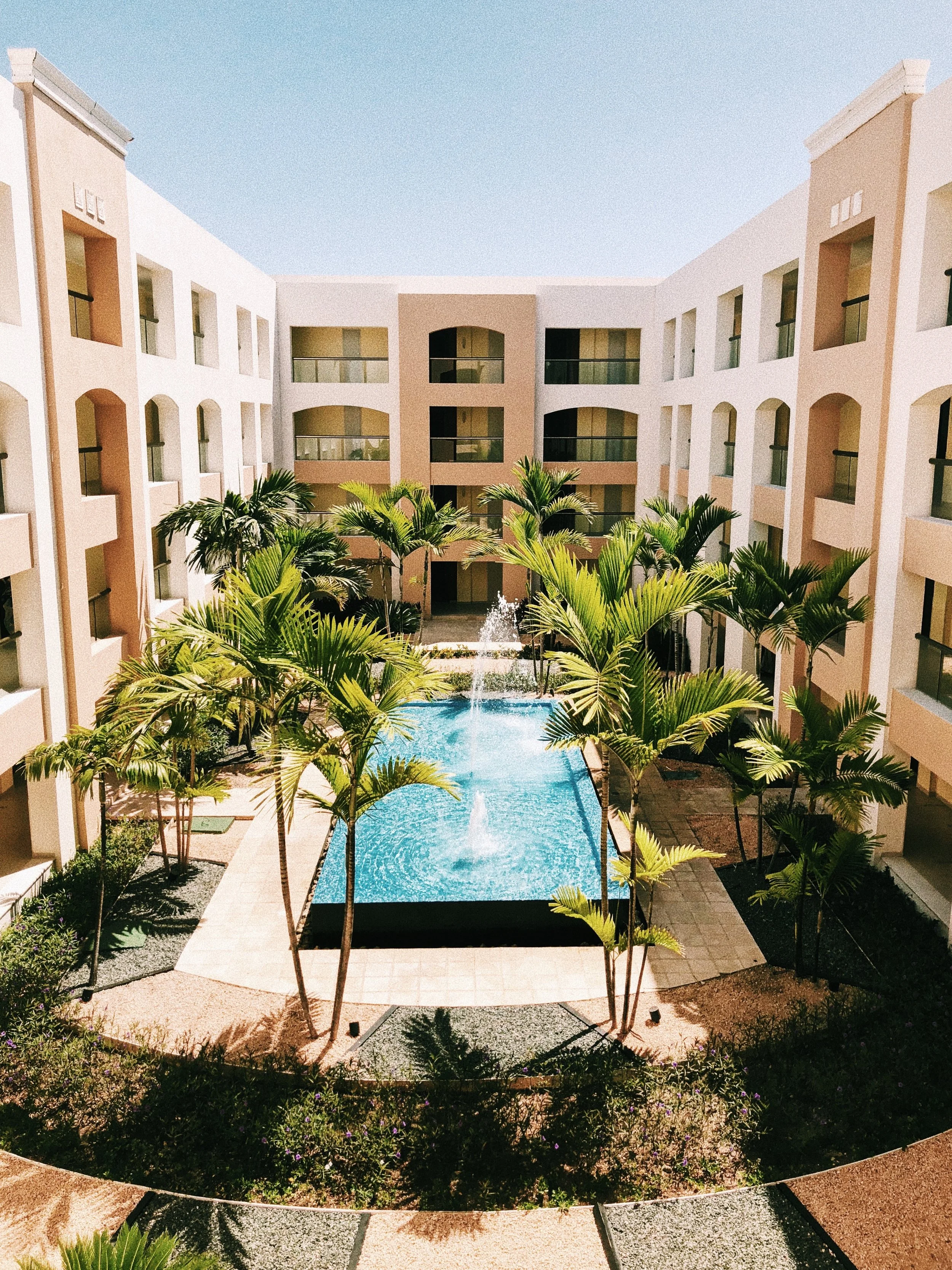 A courtyard of a hotel or apartment complex featuring a swimming pool with fountains, surrounded by palm trees, with a four-story building with balconies on each side.