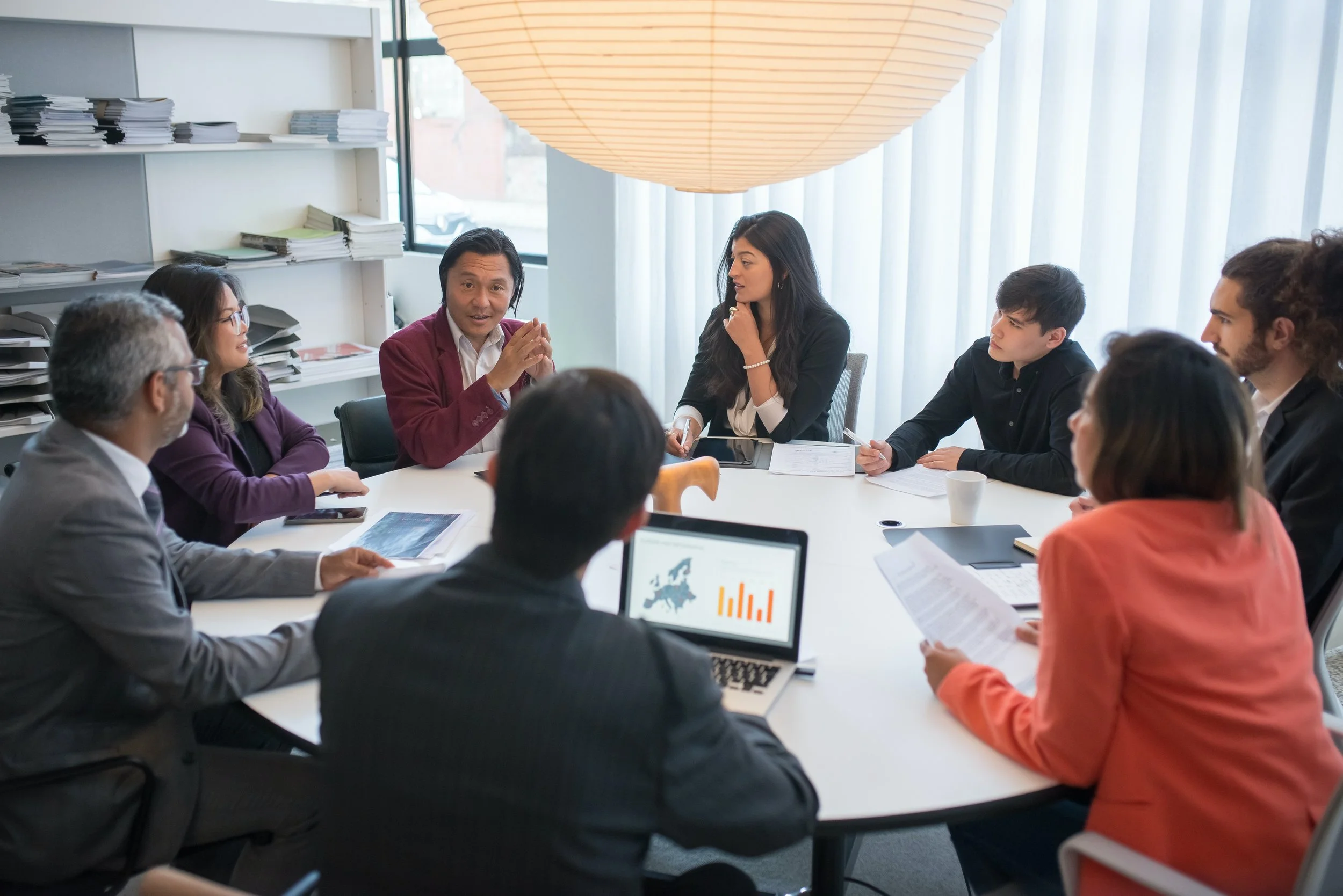Business meeting with diverse group of people sitting around a table, listening to a man in a red blazer talking, with laptops, documents, and a white coffee cup on the table, in a modern office setting.