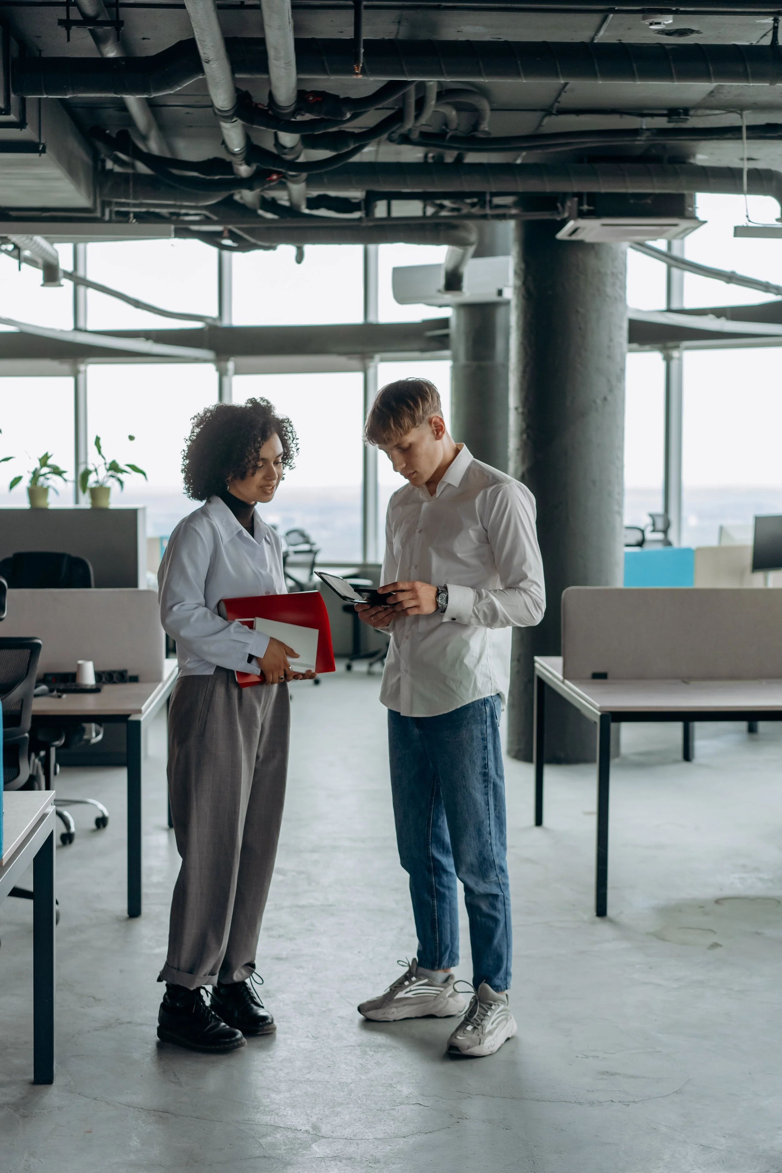 Two young professionals standing in an open office space, looking at a tablet together, with desks and large windows in the background.