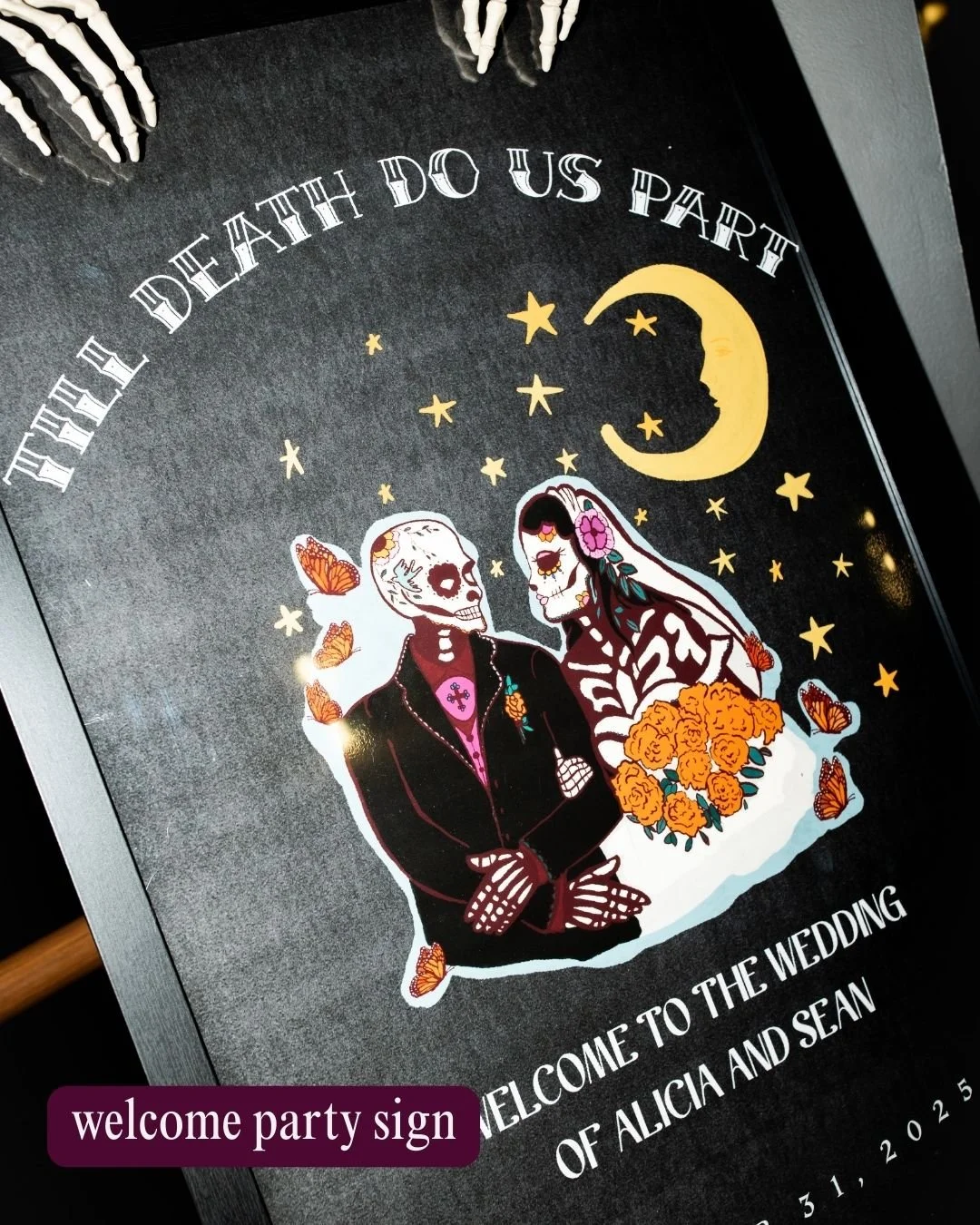 A black wedding welcome sign featuring skeleton bride and groom underneath a crescent moon and stars, with butterflies and orange flowers, celebrating a Day of the Dead themed wedding.