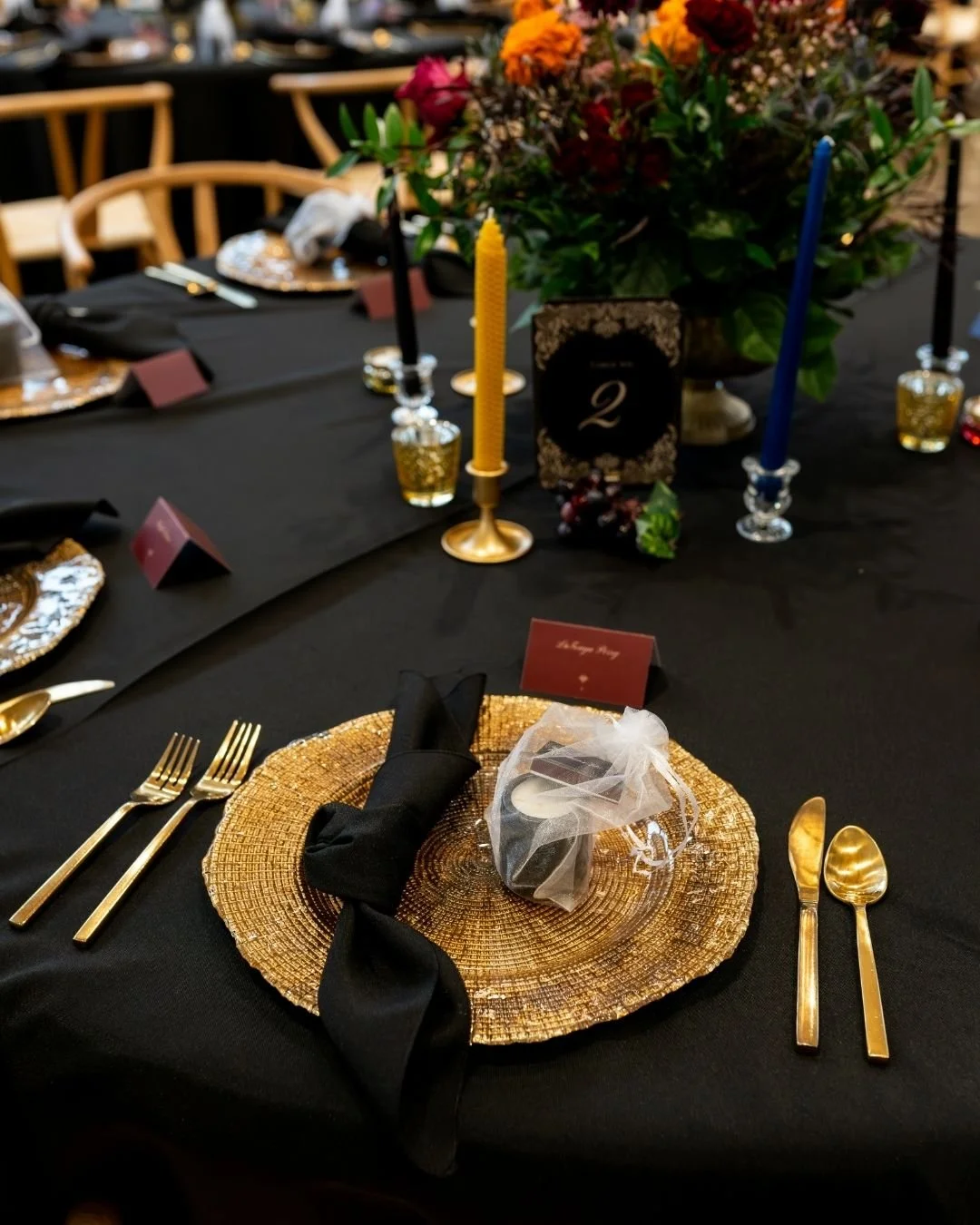 Elegant banquet table setup with gold chargers, black napkins, gold cutlery, colorful floral centerpiece, and lit candles in blue and yellow holders, indicating a formal event or celebration.