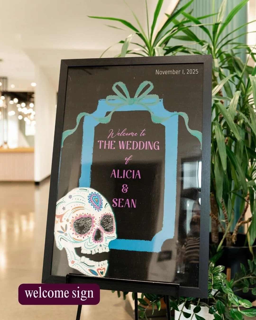 A welcome sign for a wedding featuring a large blue gift box illustration with a bow, pink text that reads "Welcome to THE WEDDING of ALICIA & SEAN," a decorated sugar skull with colorful patterns to the bottom left, and the date November 1, 2025, in the top right corner. The sign is placed next to a green plant inside a venue.