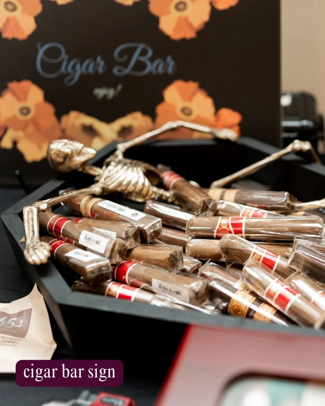 A display of individually wrapped cigarillos in a black box, with a skeleton decoration on top, and a sign in the background reading 'Cigar Bar' with orange flower designs.