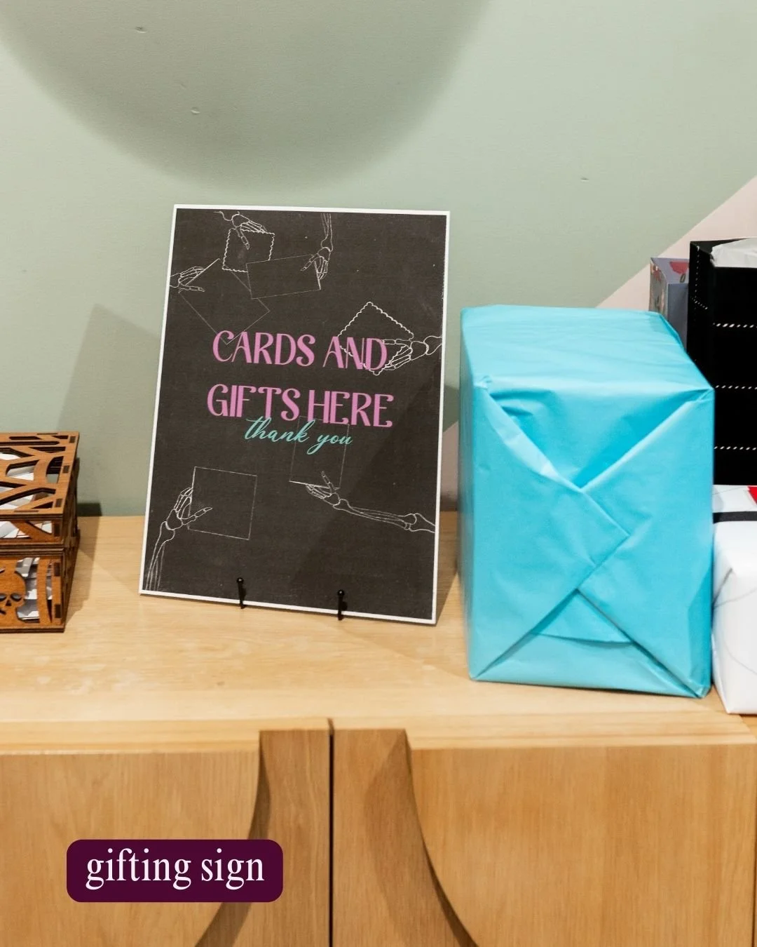 Gifting sign on a wooden table with cards and gifts nearby, including a large blue wrapped gift, in an indoor setting.