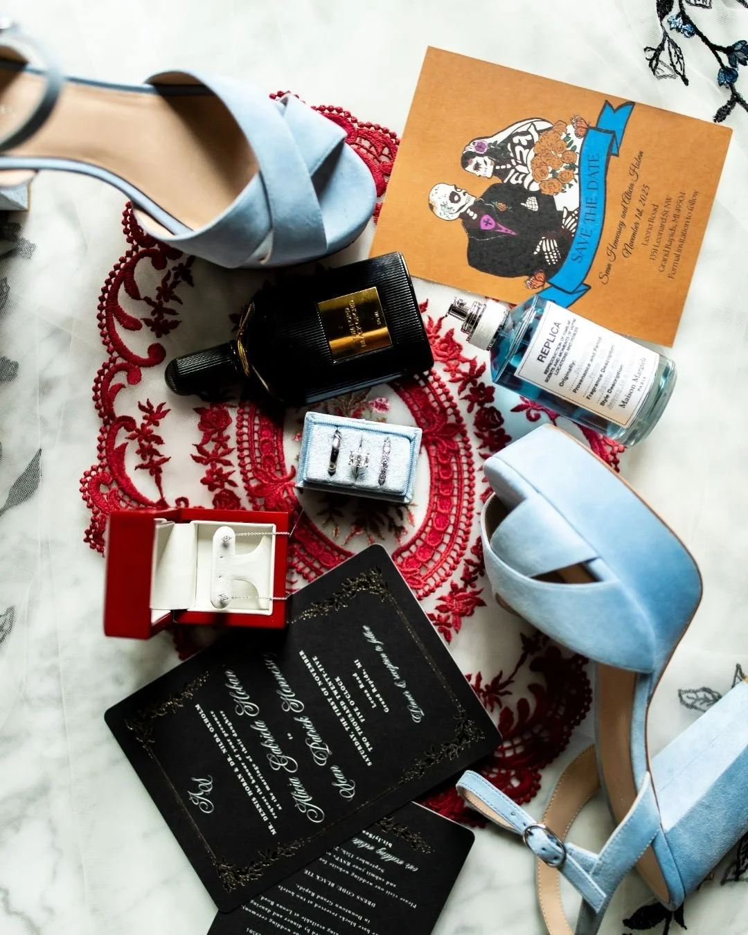 A collection of wedding accessories and invitations displayed on a red embroidered cloth, including light blue high-heeled shoes, a black perfume bottle, a small jewelry box with earrings, a red jewelry box with earrings, wedding invitation cards, a bottle of Repluxa disinfectant, and a save-the-date card with a skeleton couple illustration.