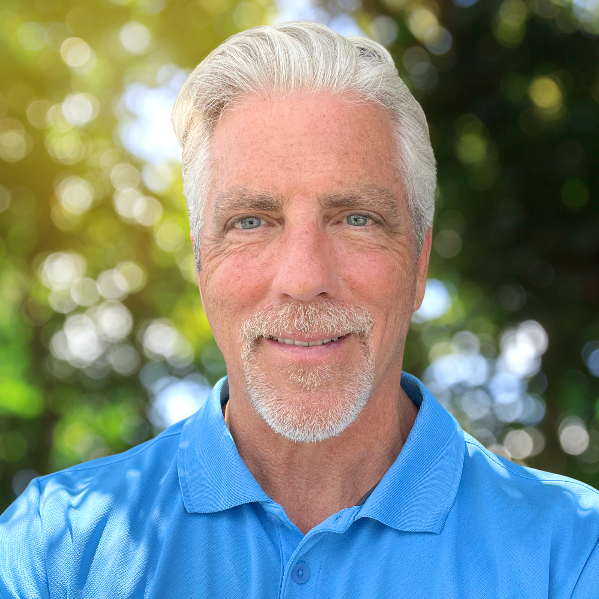 Portrait of an older man with gray hair and a beard, wearing a blue collared shirt, smiling outdoors with blurred green foliage in the background.