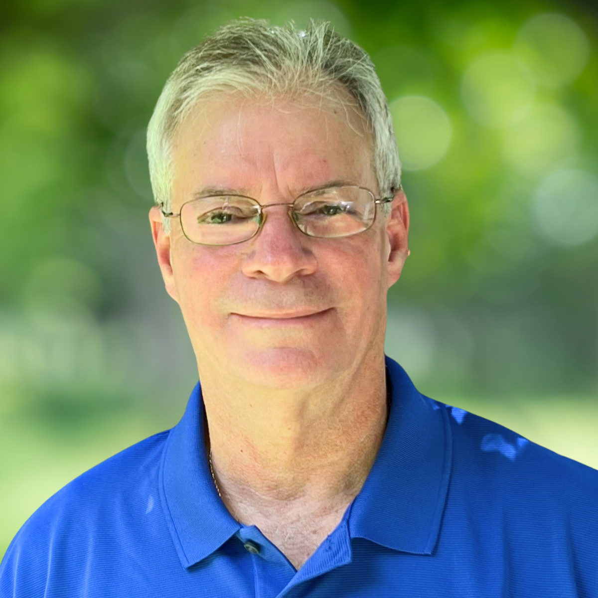 A middle-aged man with gray hair, glasses, and a slight smile, wearing a blue collared shirt, standing outdoors with a blurred green background.
