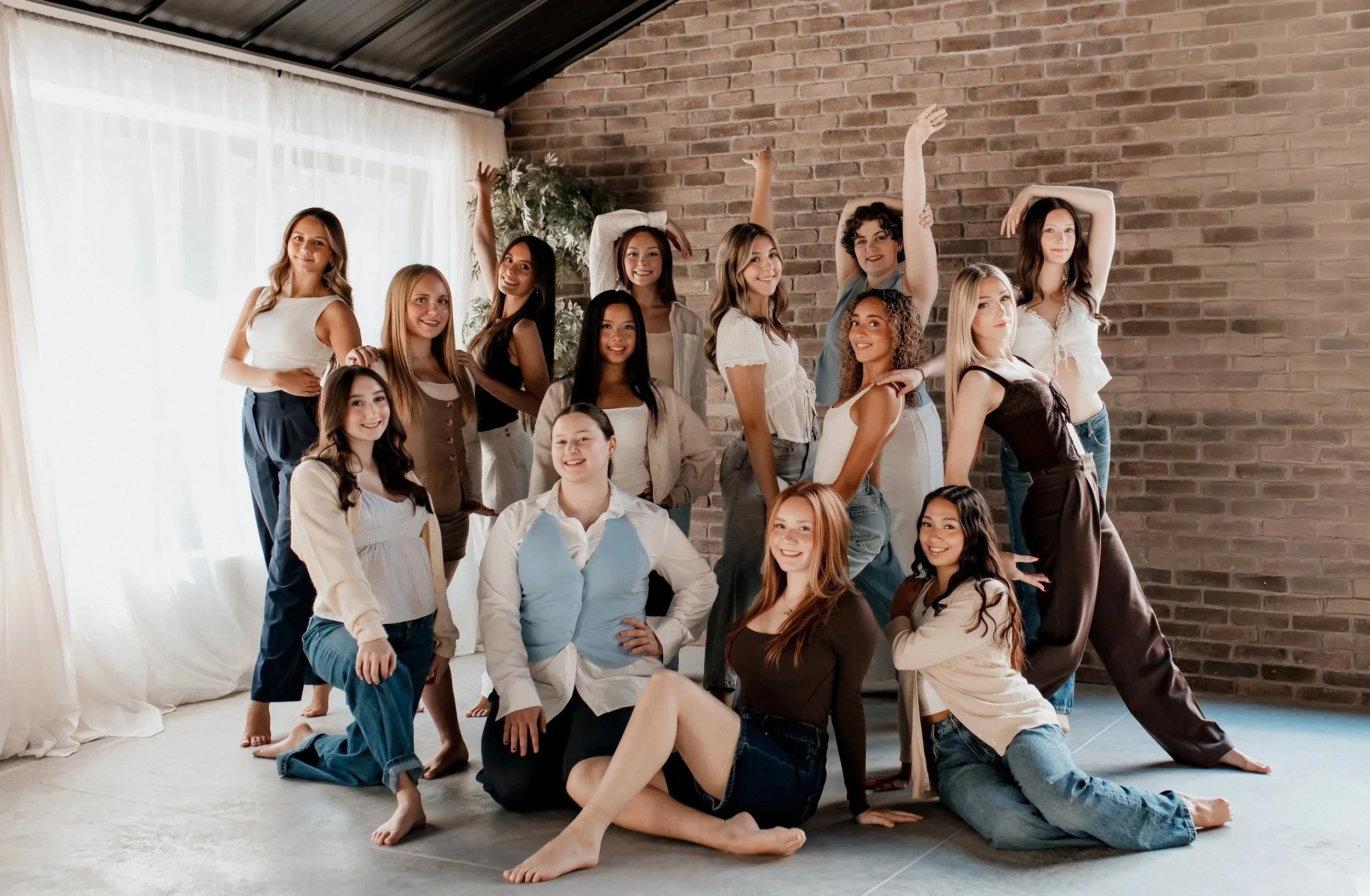 A group of fifteen young women posing indoors in front of a brick wall, with some kneeling, sitting, and standing, smiling and looking at the camera.