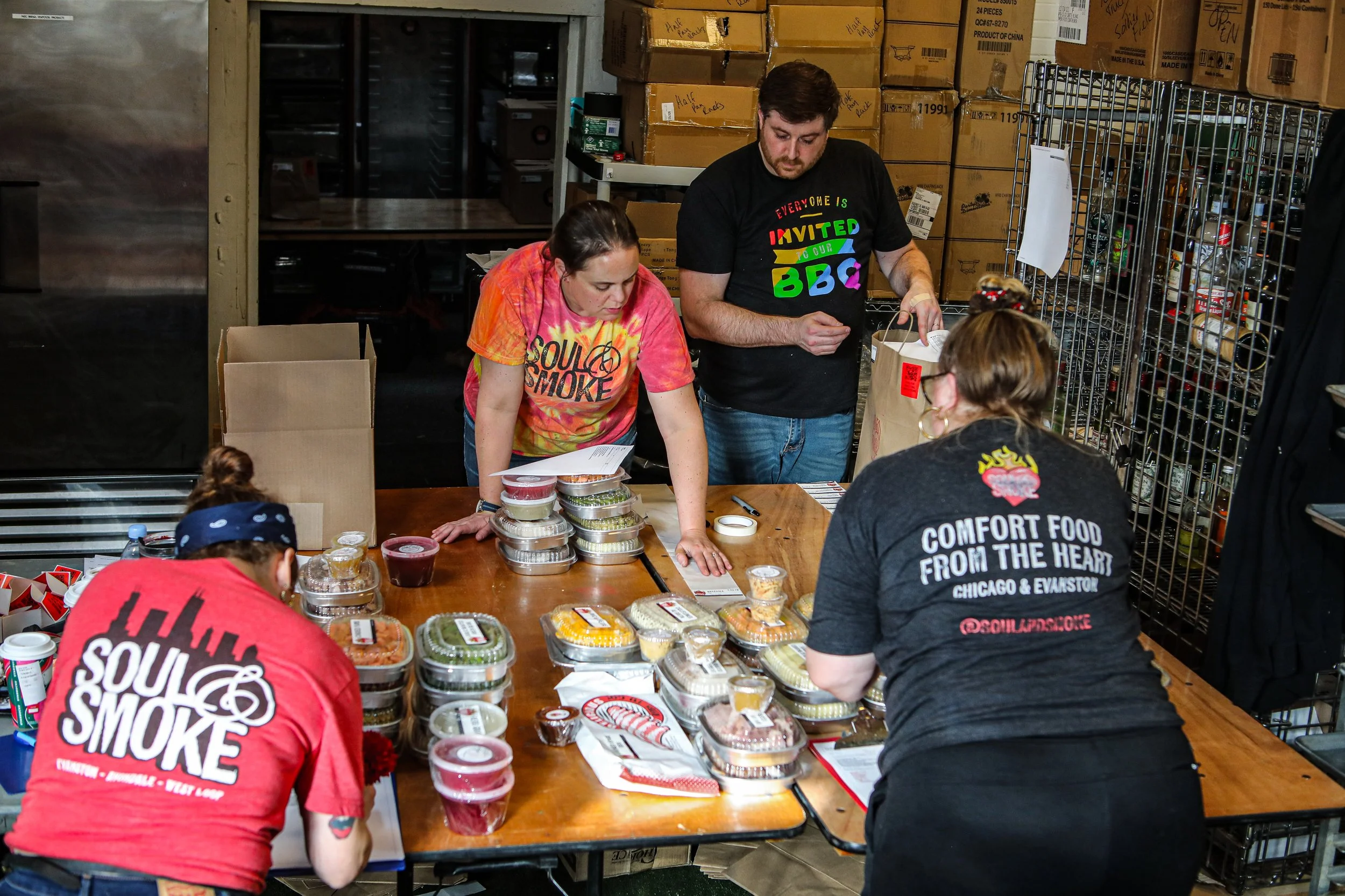 Four people are gathered around a table filled with food containers, at a warehouse with boxes and metal racks in the background.