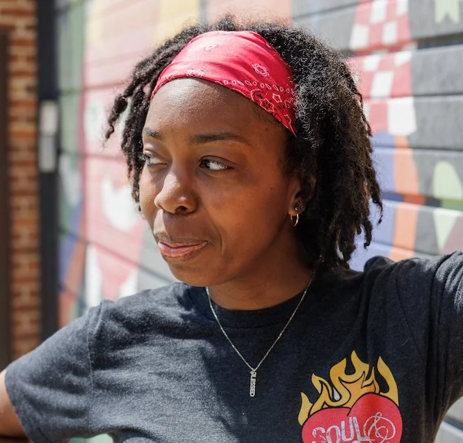 A woman with medium brown skin and curly hair, wearing a red bandana, black T-shirt with a heart and flames design, and a small necklace, standing in front of a colorful graffiti wall.
