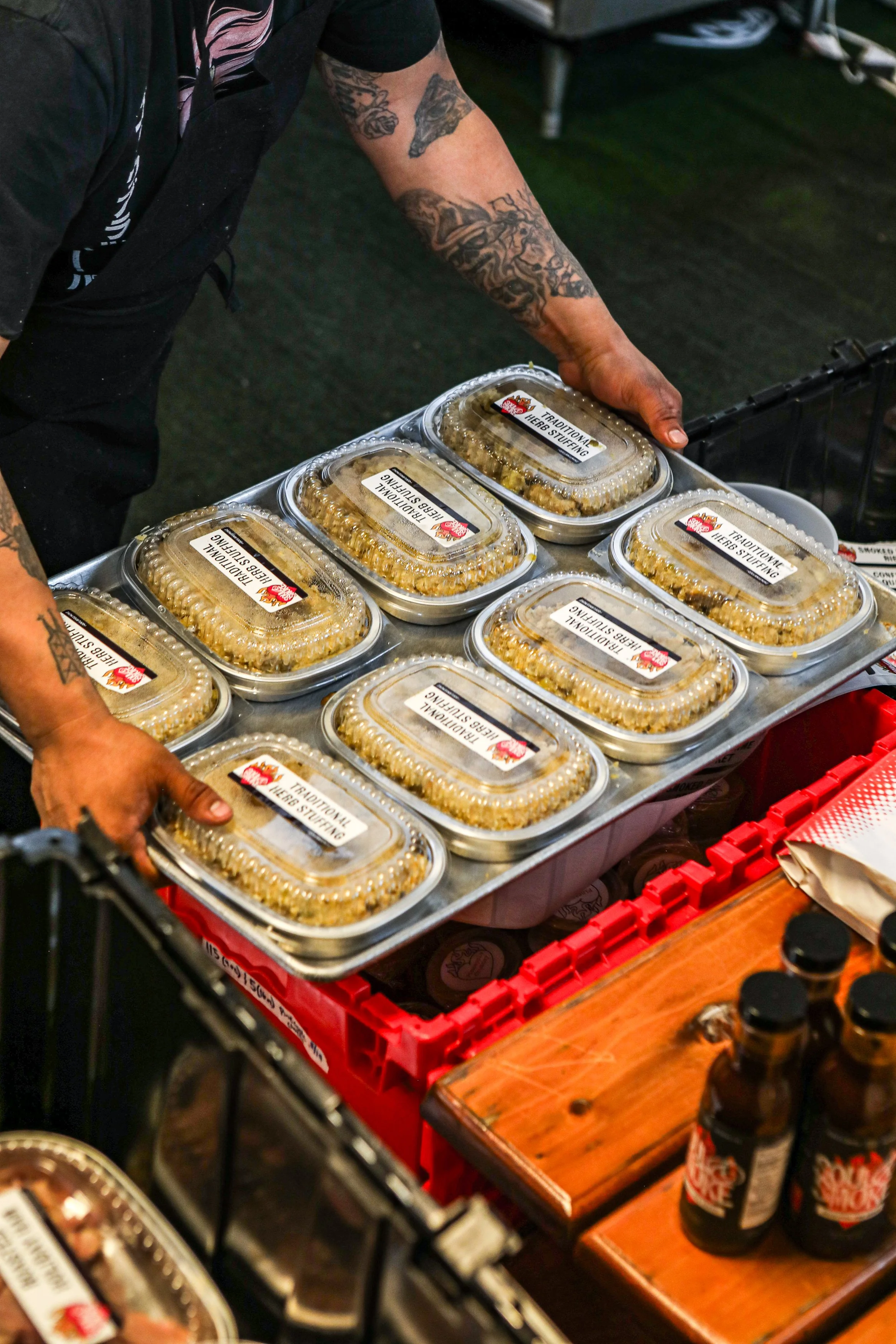 A person with tattoos on their arms is handling a tray of pre-packaged herb stuffing from a food stand or store. The tray contains multiple serving containers with clear lids labeled 'Traditional Herbs Stuffing.' There are also bottles of sauce or dr