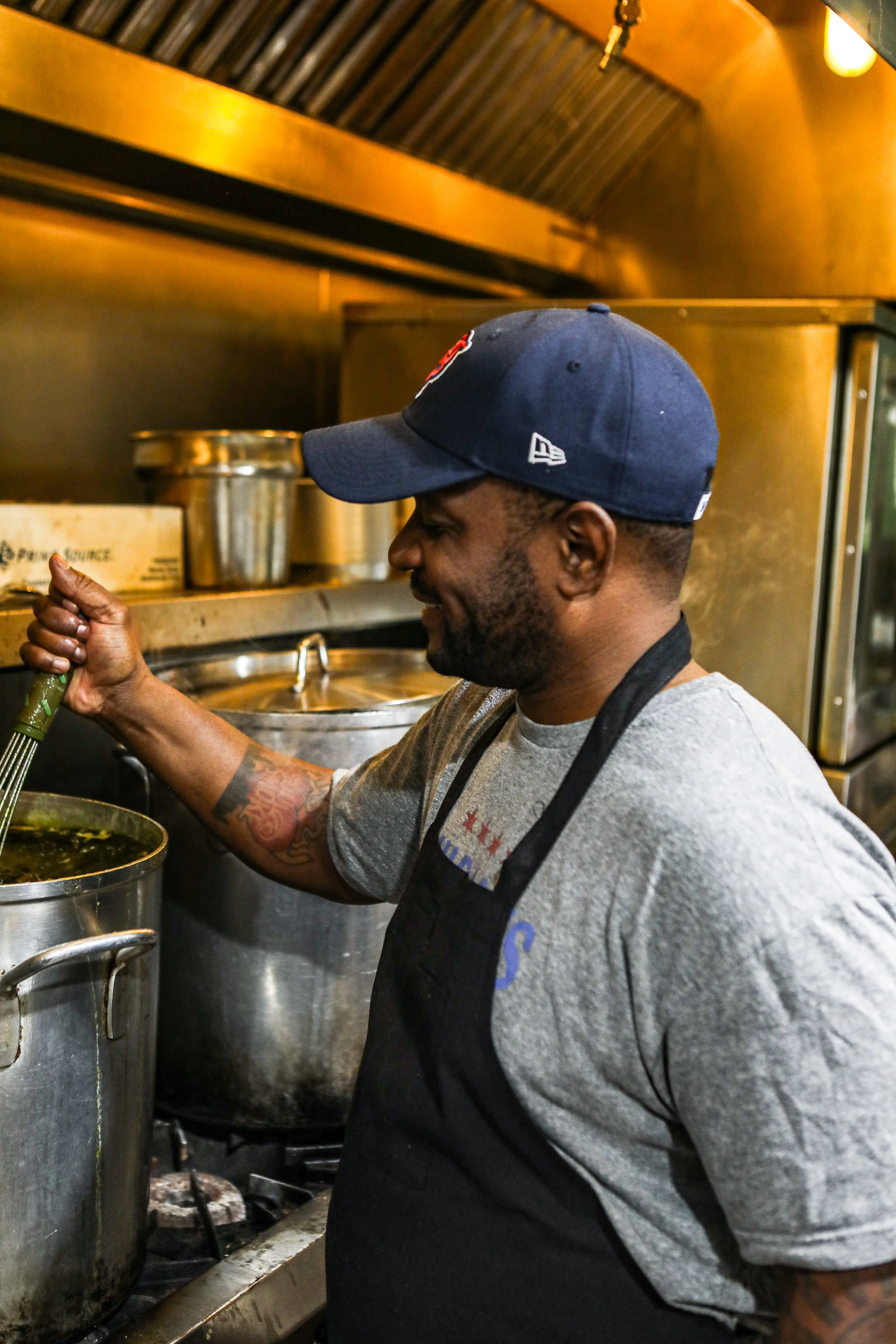 A man cooking in a commercial kitchen, wearing a baseball cap and a black apron, stirring a large pot of soup or stew.