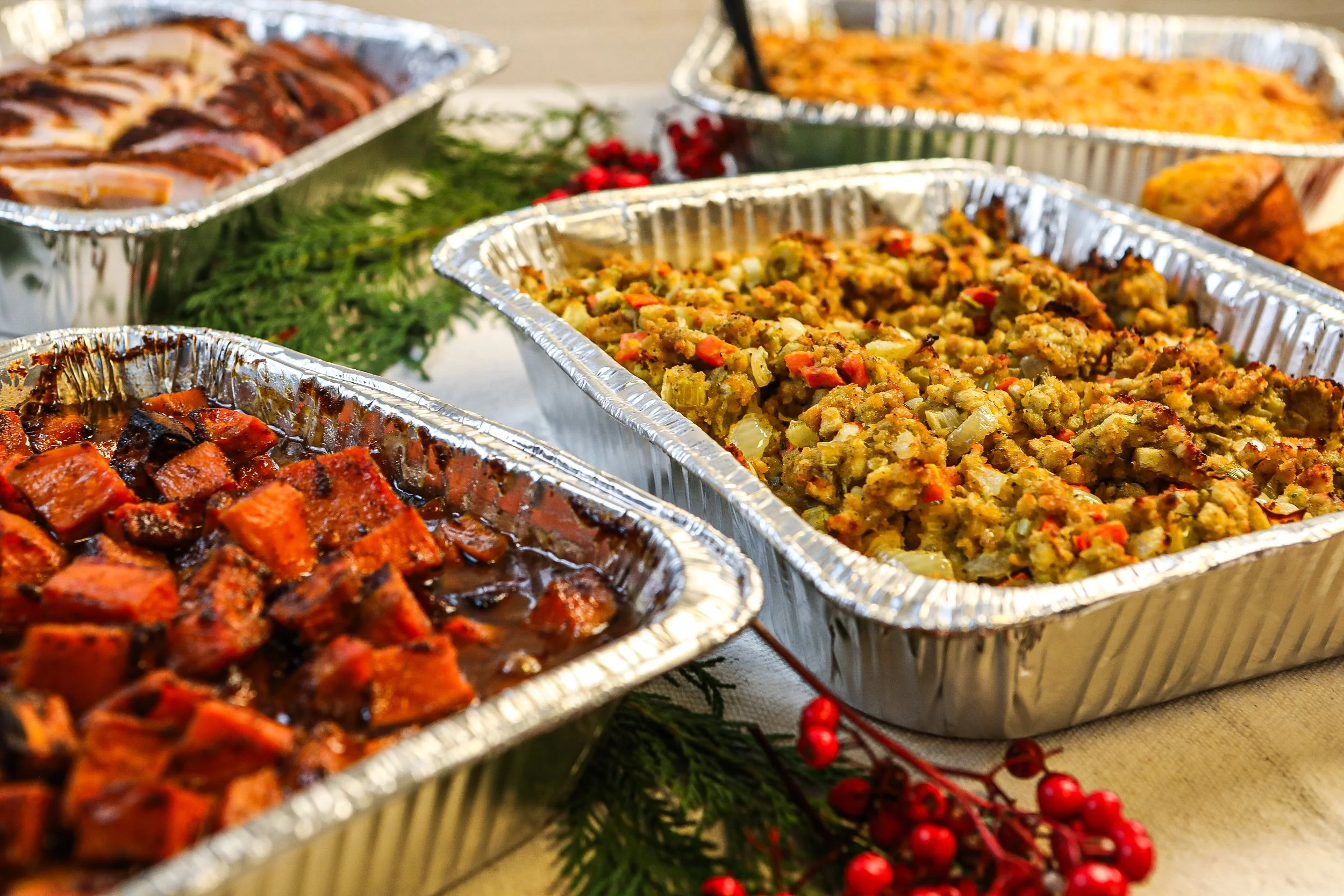 Various dishes of food in aluminum trays on a table, including sweet potatoes, stuffing, and glazed carrots, decorated with holiday greenery and red berries.