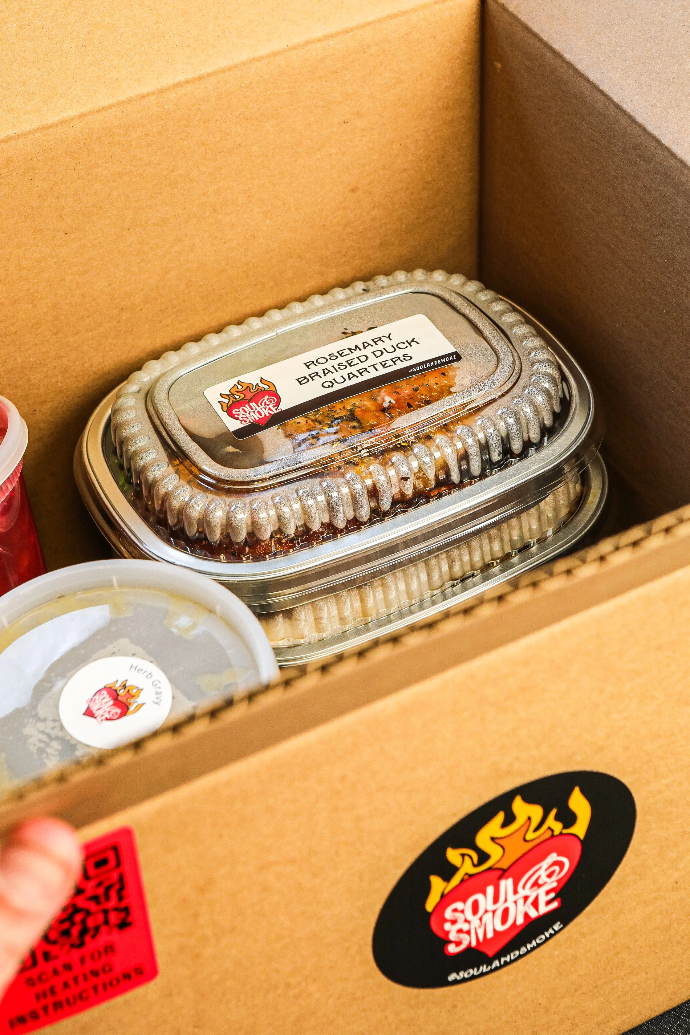 A cardboard delivery box containing containers of smoked food, including a tray labeled Rosemary Braised Duck Quarters