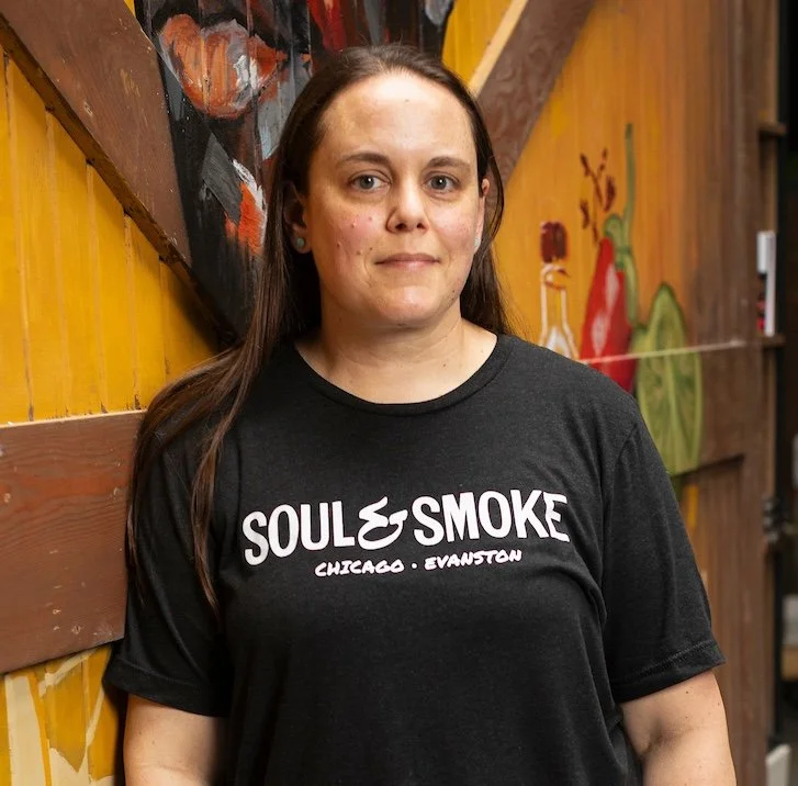 A young woman smiling outdoors in front of a colorful mural, wearing a red t-shirt with a flame and the word 'Souls' on it.