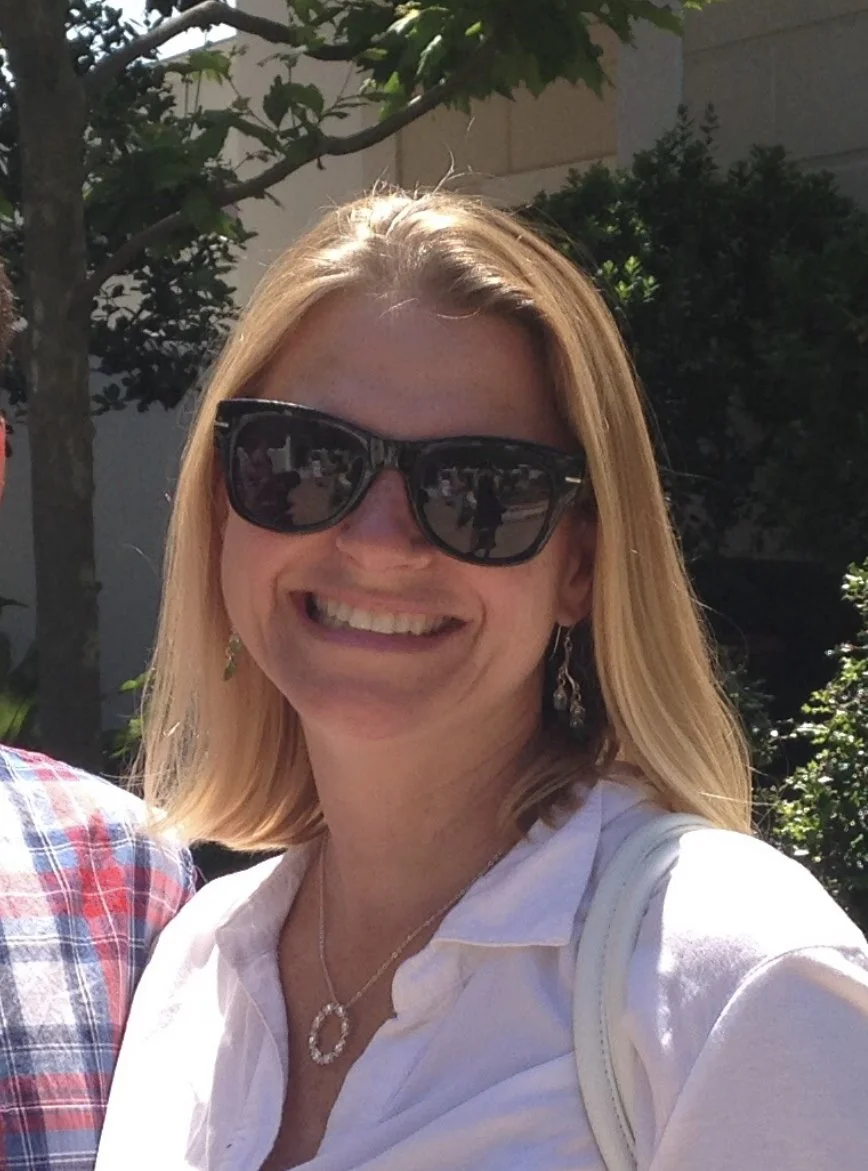 A smiling woman with blonde hair wearing large black sunglasses, a white shirt, and earrings is outdoors with greenery and a building in the background.