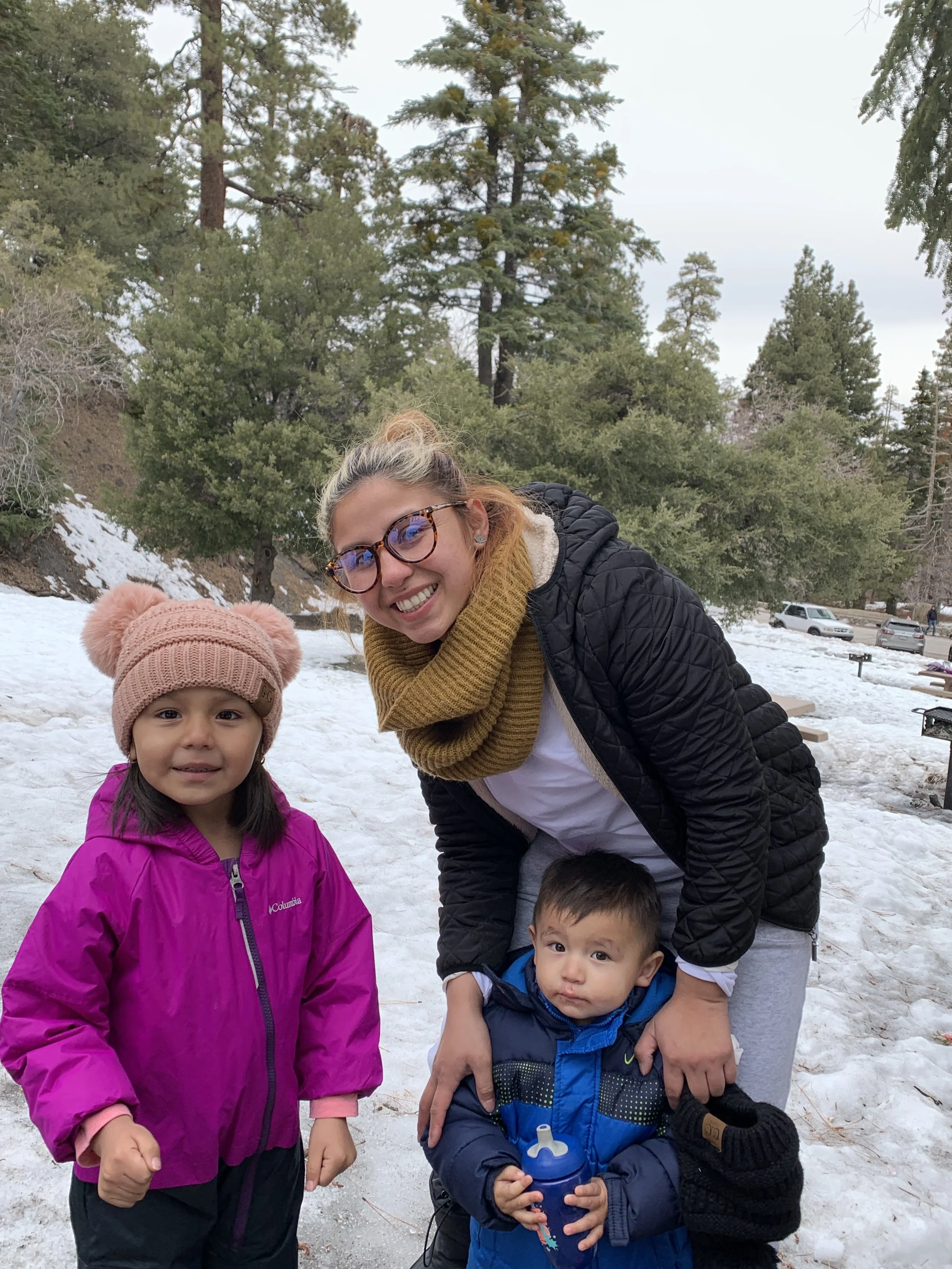 A smiling woman with glasses and a brown scarf posing outdoors with two children in a snowy, forested area. The girl is wearing a pink jacket and a pink knit hat, while the young boy is dressed in a blue jacket and holding a water bottle. Pine trees and parked cars are visible in the background.