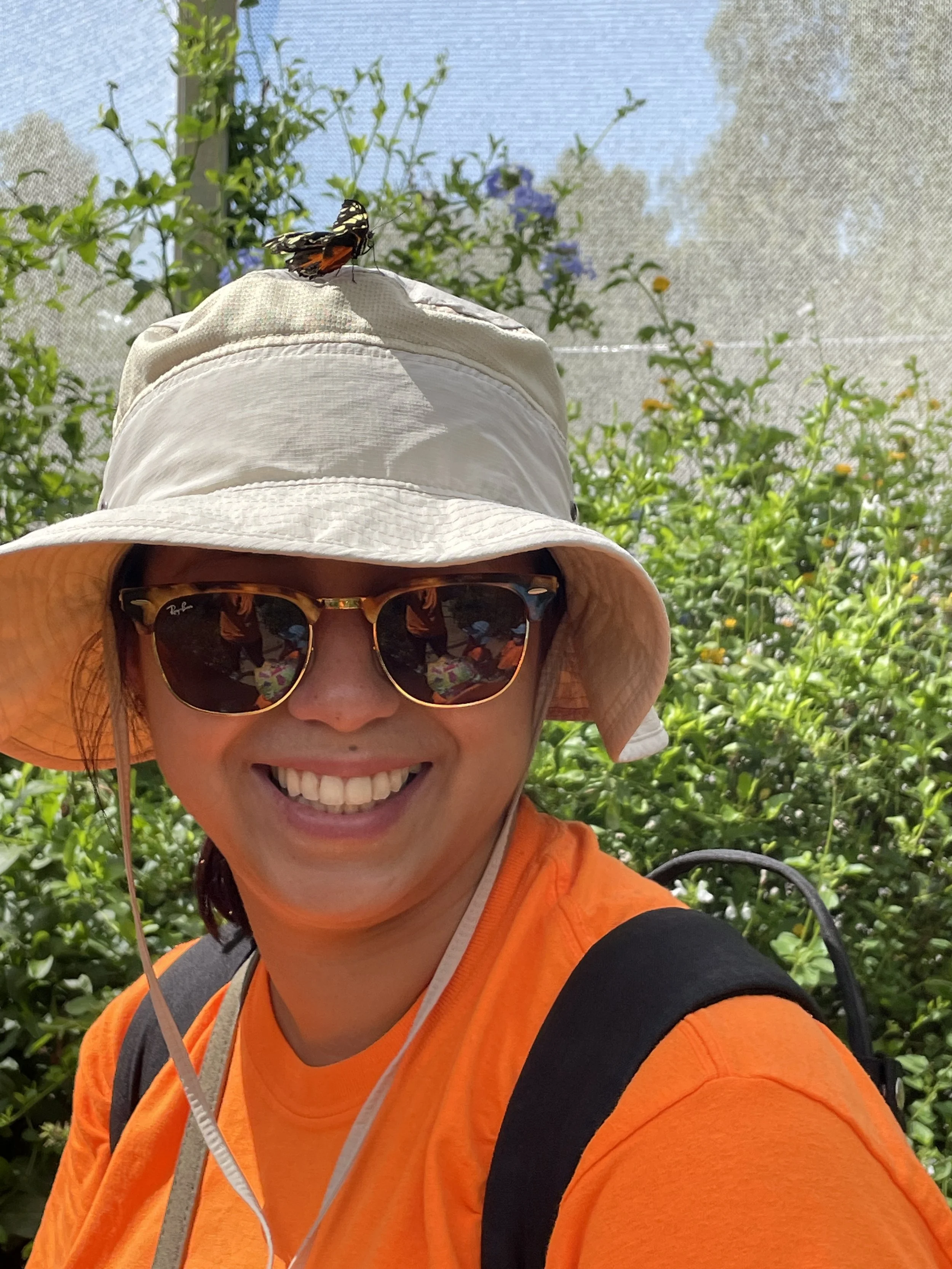 A woman smiling outdoors in front of green bushes, wearing sunglasses, an orange shirt, a beige wide-brim hat with a butterfly perched on top, and a black backpack.