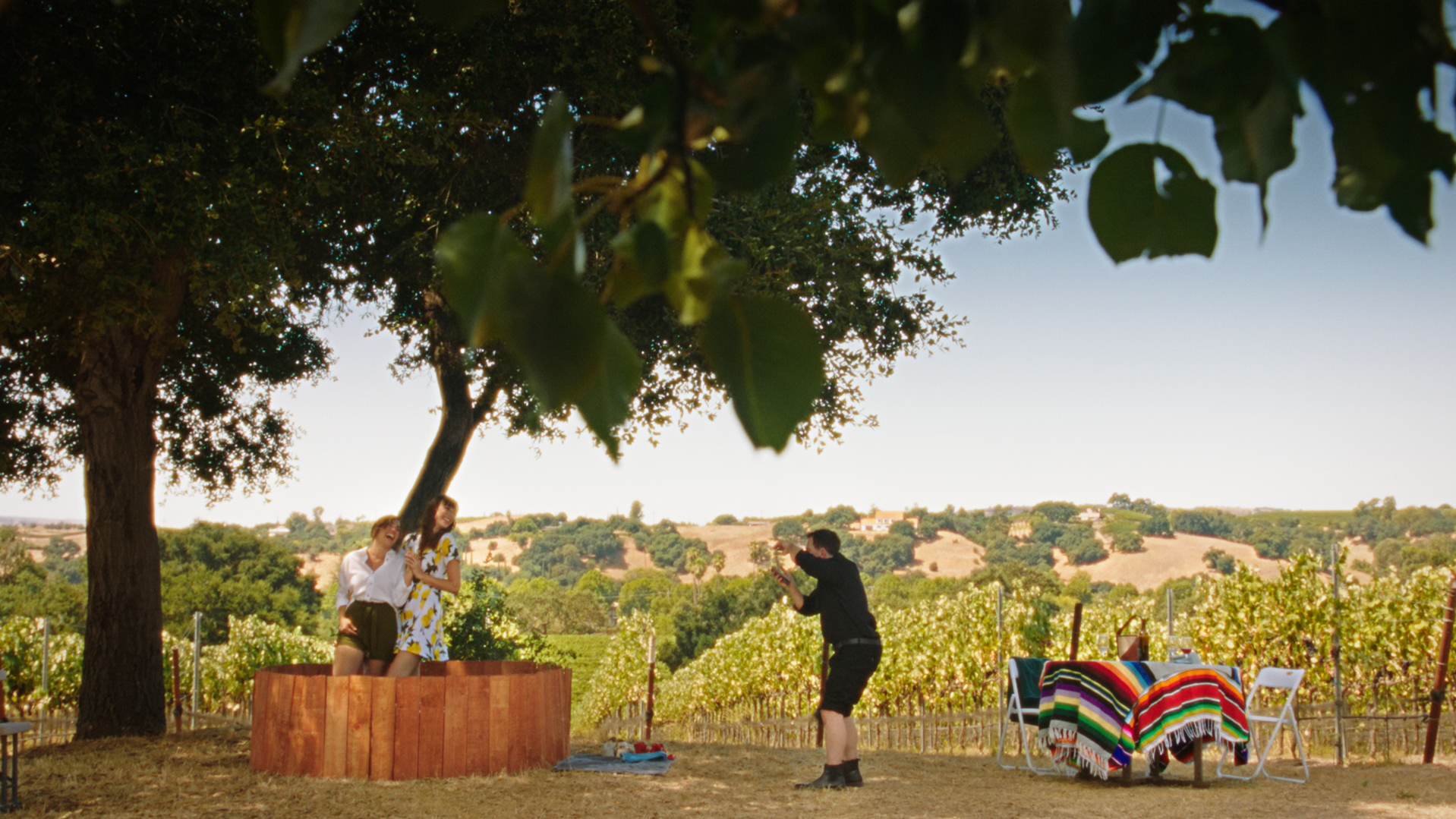 People taking photos of two women standing inside a wooden border, with a photographer capturing the moment, in a vineyard setting with a vineyard, trees, and rolling hills in the background.