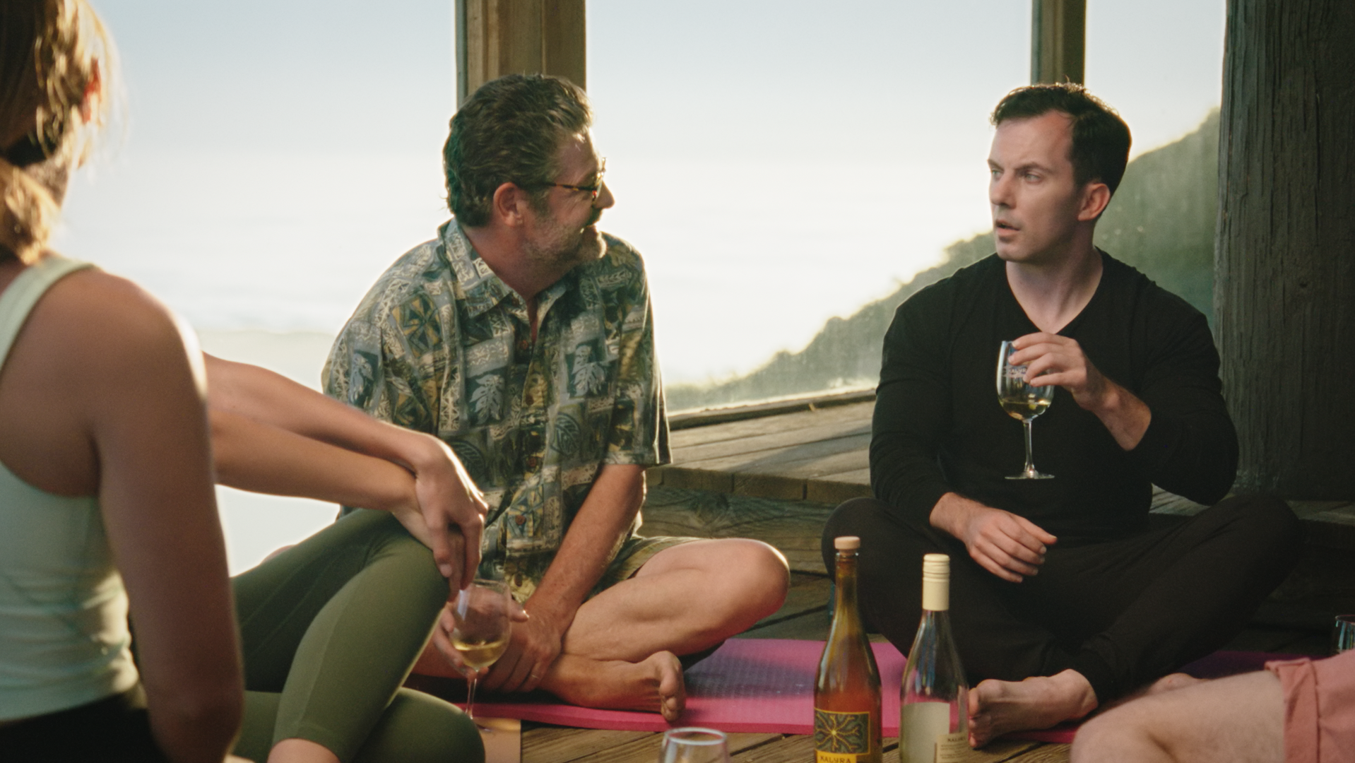 Group of people sitting on a wooden floor with yoga mats, having drinks, by a window showing an outdoor view with trees and the ocean at sunset.
