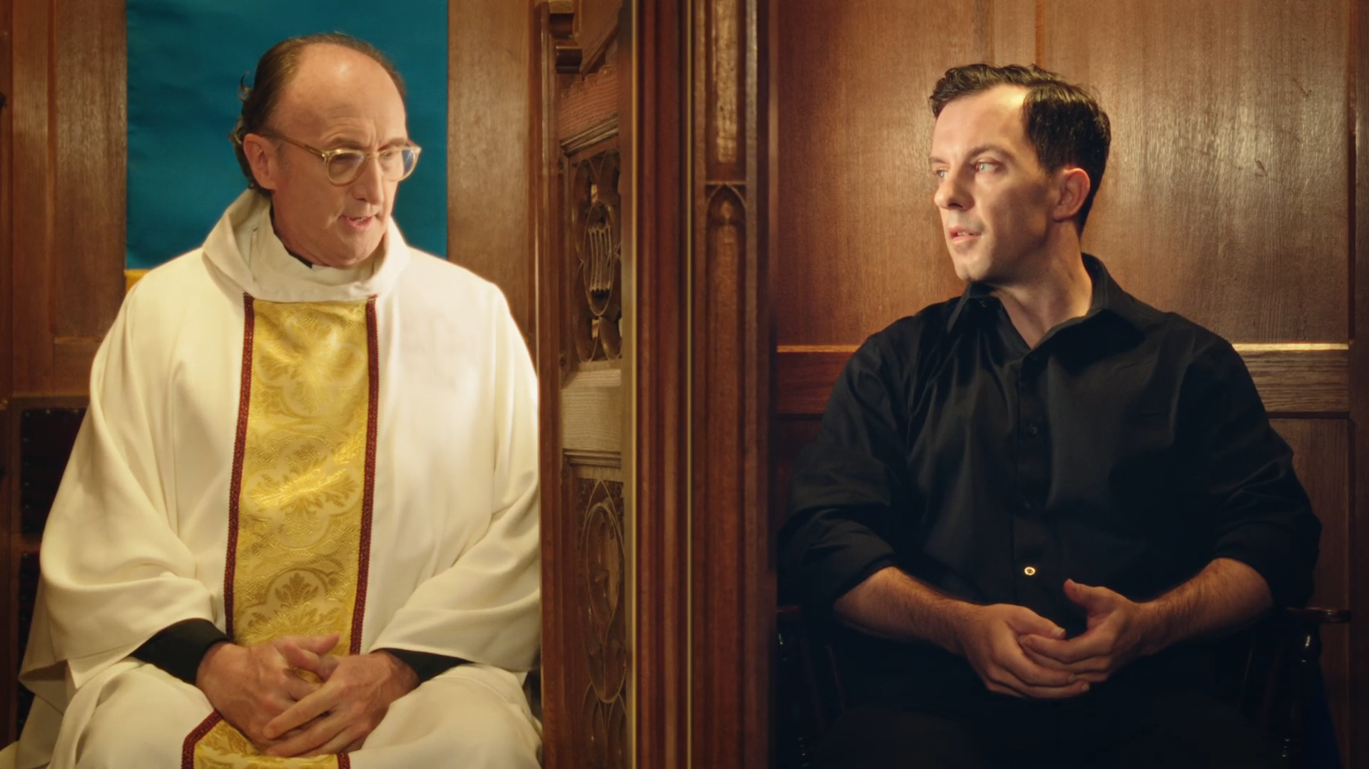 A priest in white and gold robes in conversation with a man in black seated in a wooden-paneled room.