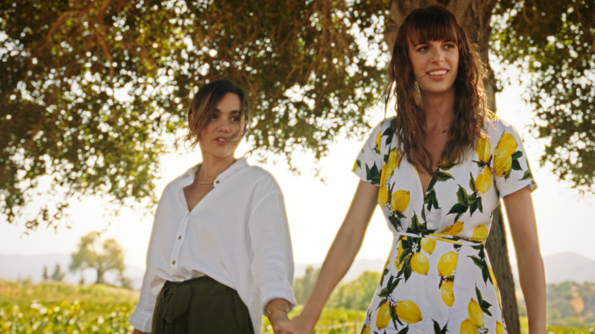 Two women holding hands outdoors, one wearing a white blouse and the other a lemon-patterned dress, standing beneath a large tree with a scenic background of fields and hills.