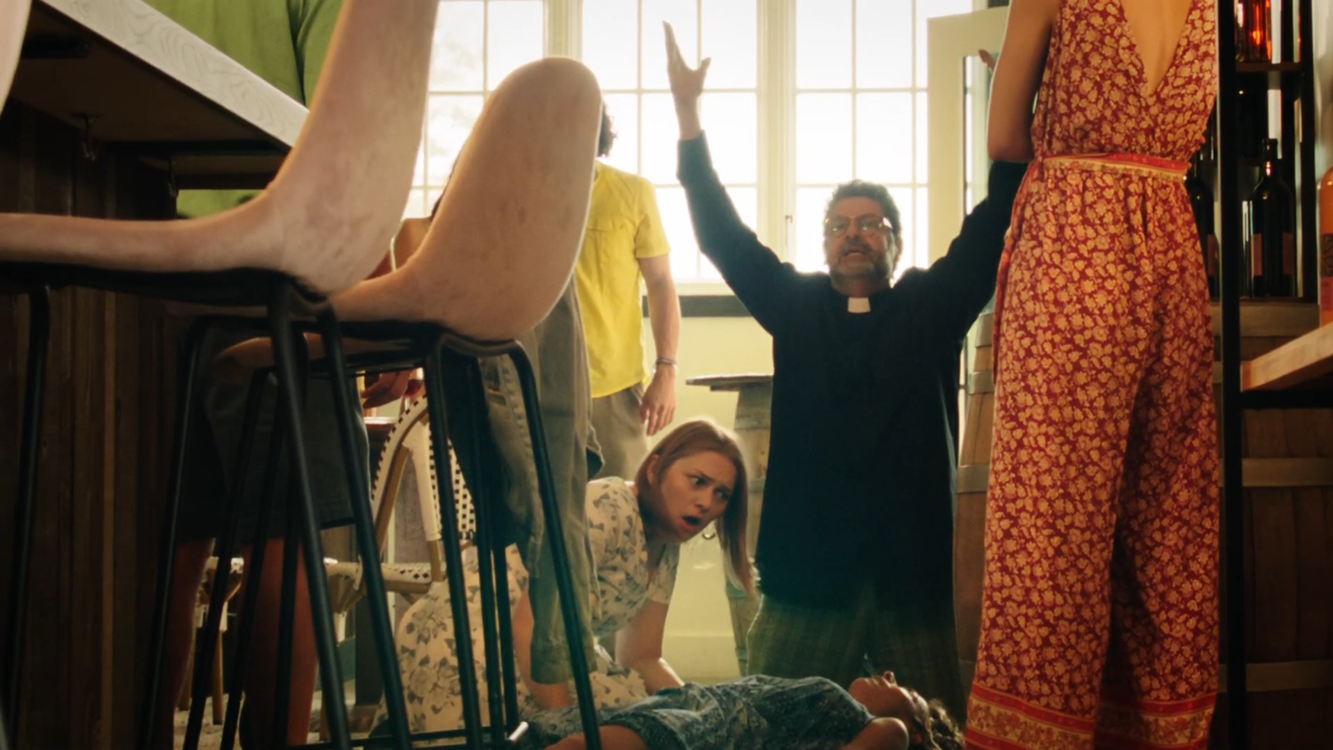 People in a room reacting in shock or surprise, with a woman lying on the floor and others bending or kneeling around her, a man dressed as a priest has arms raised, and a woman in a red and orange patterned dress is standing.