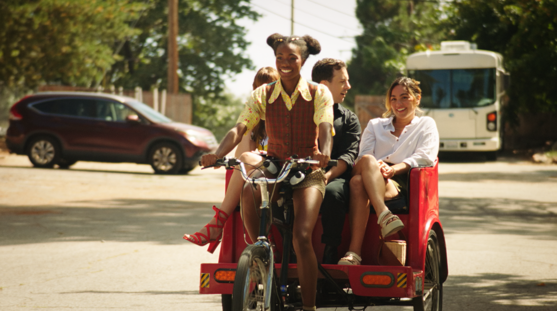 Four young people enjoying a ride on a tricycle with a cargo seat, riding on a sunny street lined with trees, cars, and an RV in the background. All are smiling and dressed casually.