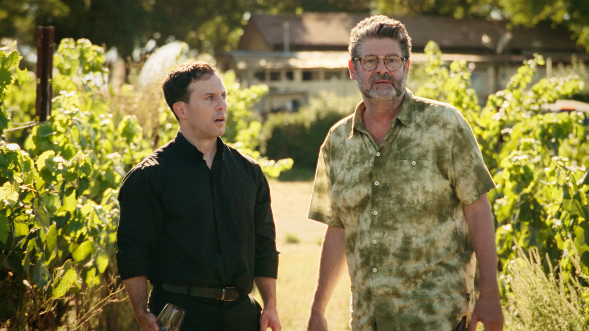 Two men walking through a vineyard on a sunny day, one holding a glass of wine, with trees and a house in the background.