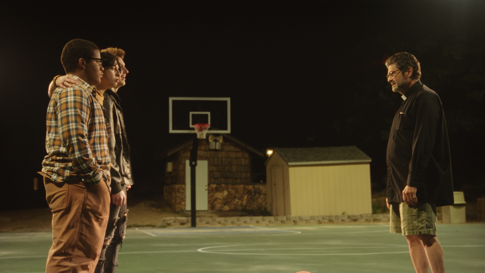 Three teenagers standing on a basketball court at night facing a man dressed as a priest. The court has a basketball hoop, and there are small buildings in the background.