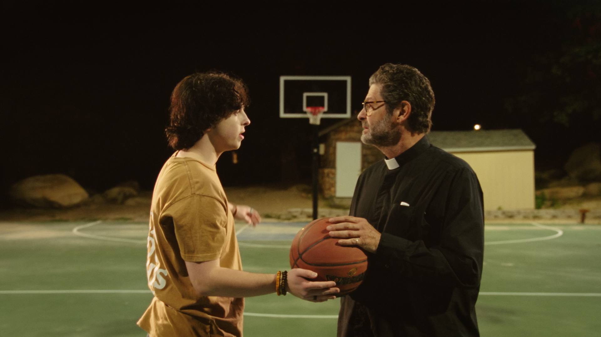 A young person and a priest are standing on an outdoor basketball court at night, engaging in a conversation while the priest holds a basketball.