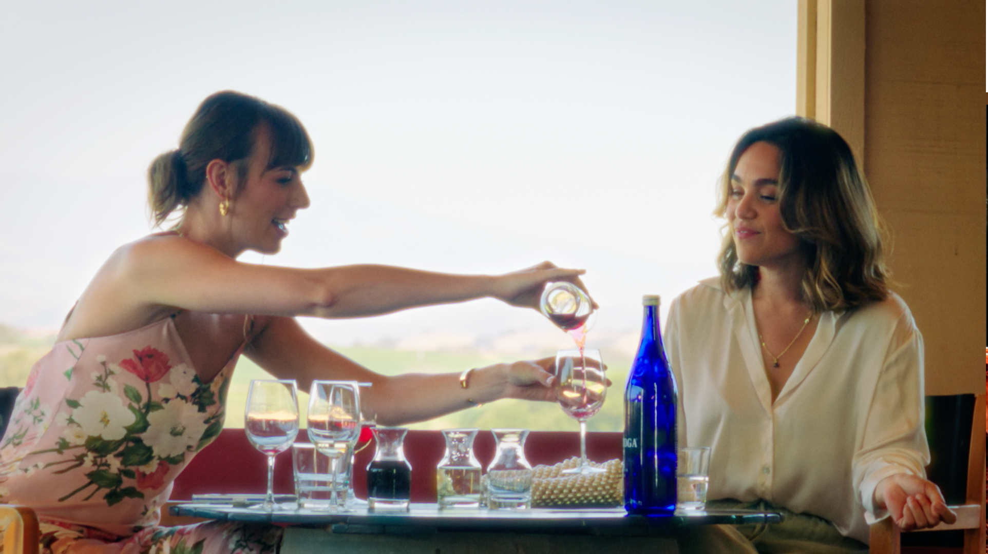Two women sitting at a table, one pouring wine into a glass for the other, with multiple glasses and bottles on the table, in a well-lit room with a large window showing an outdoor view.