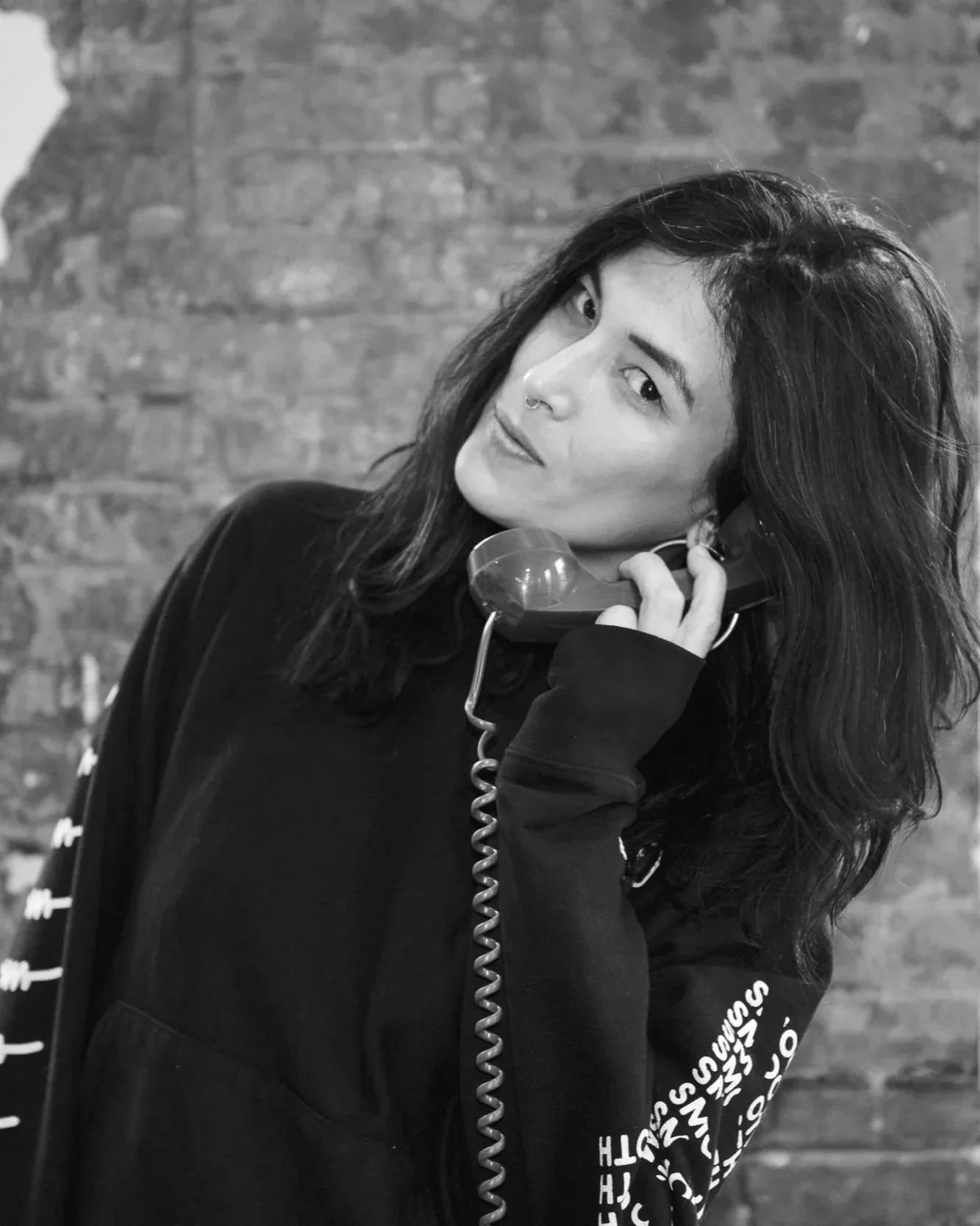 A woman with dark wavy hair, wearing a dark hoodie, holding a vintage telephone receiver to her ear, with a brick wall background, in black and white.
