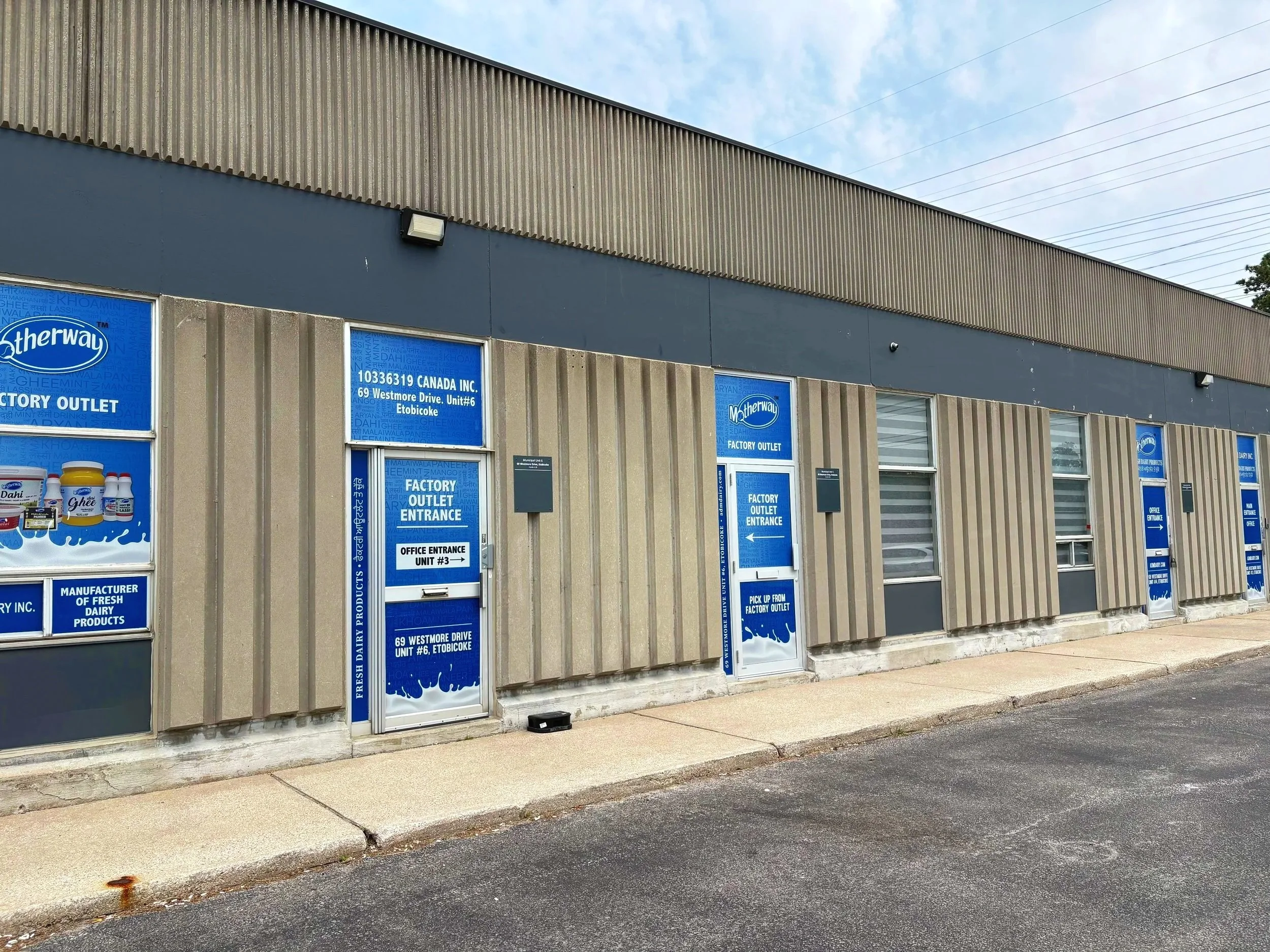 Photo of the exterior of a factory outlet building with blue signs on the windows, located at 69 Westmore Drive, Unit 6, in Etobicoke. The building has a gray, textured facade and a corrugated metal upper section. The sky is partly cloudy, and there is a sidewalk and street curb in the foreground.