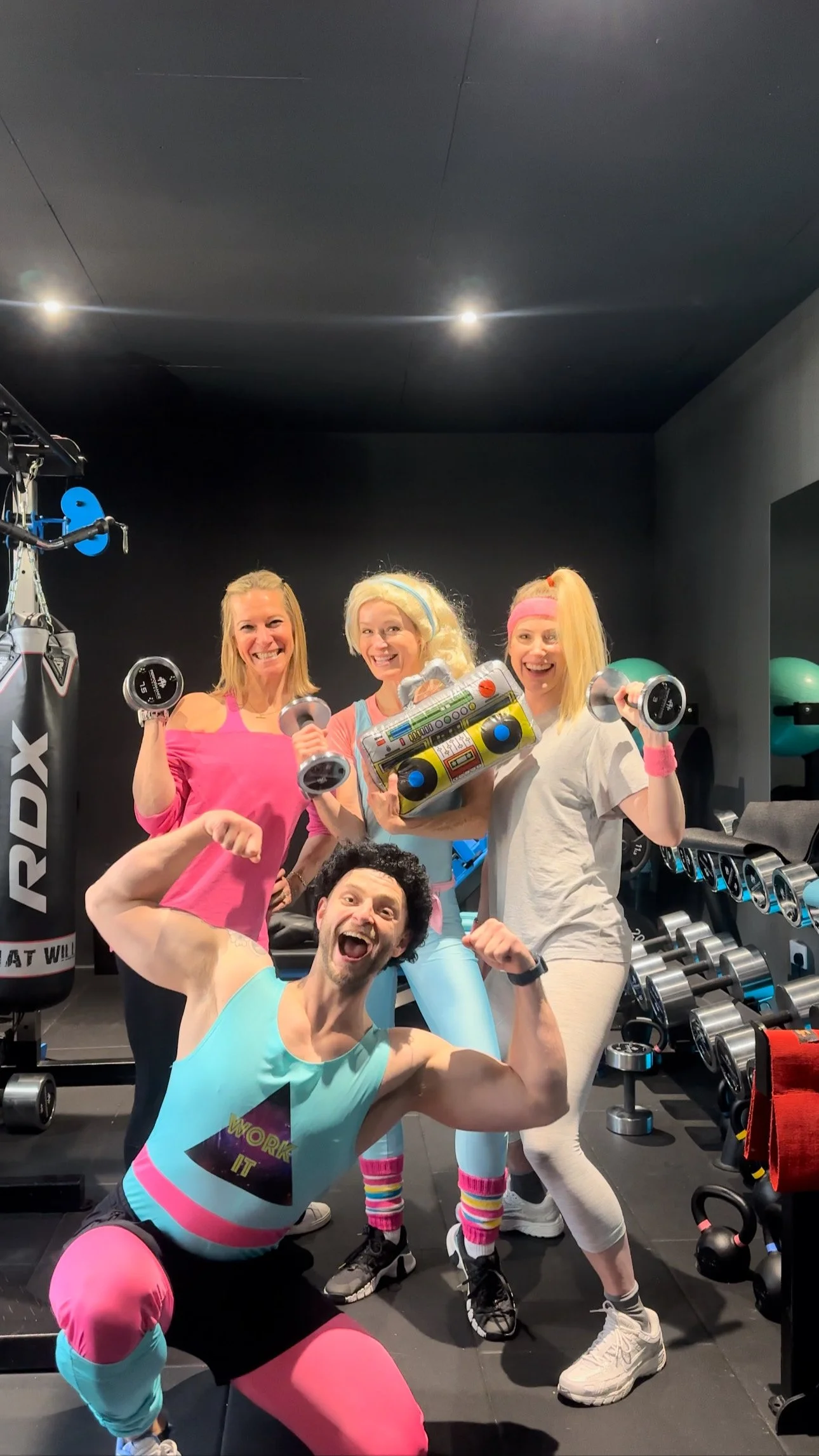 Group of five people in workout attire at a gym, smiling and posing with dumbbells and a boombox, surrounded by exercise equipment and a punching bag.